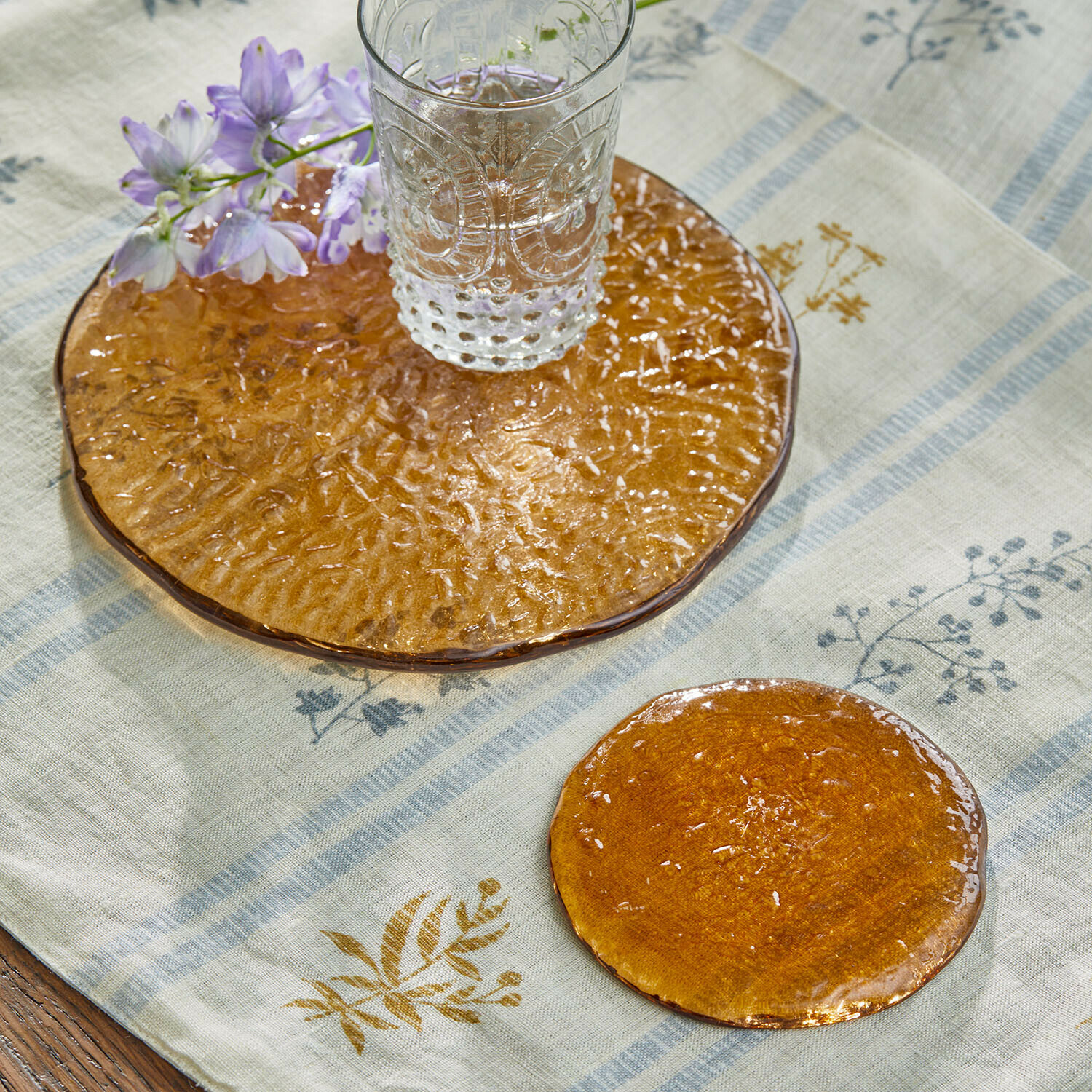 Coppia di sottobicchieri Montelisse Bicchiere di vetro trasparente su sottobicchiere in vetro arancione con fiore su tovaglia di lino