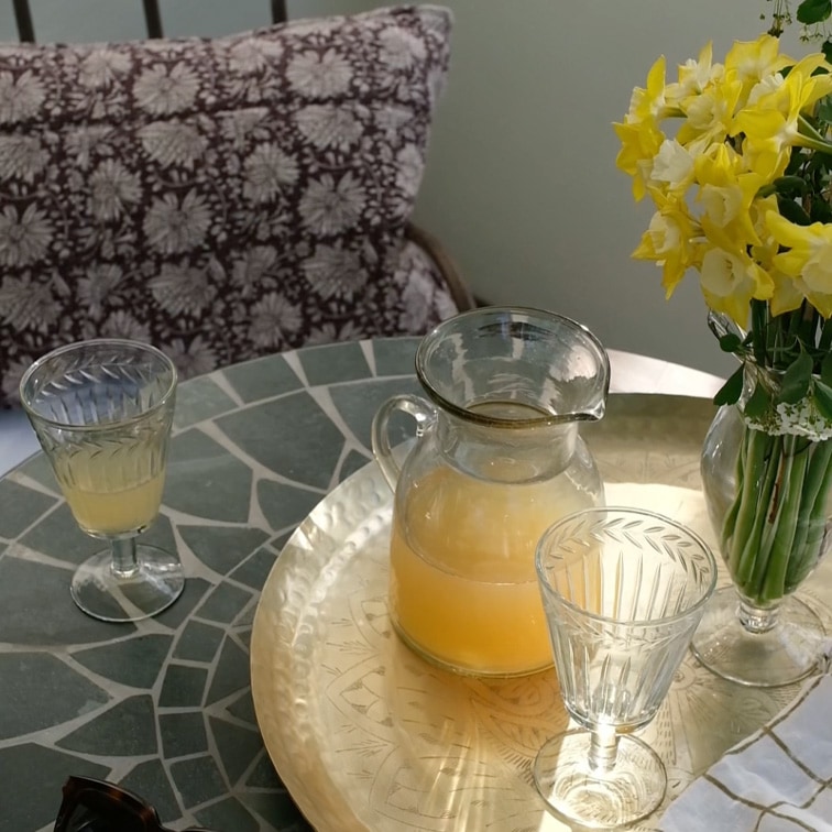 Caraffa di vetro con bevanda gialla e due bicchieri su tavolo in marmo con fiori in vaso