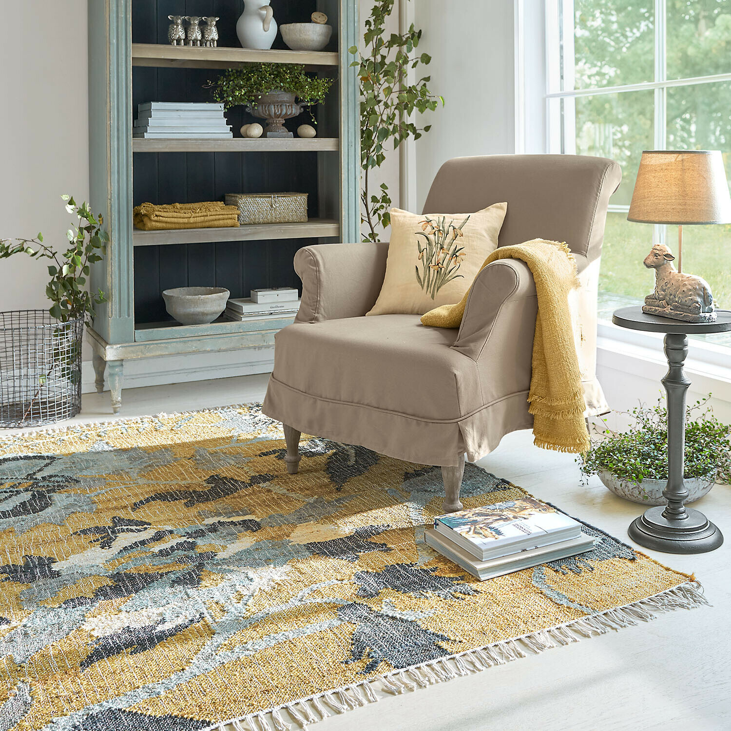 Tappeto Ornelless Poltrona beige con cuscino e coperta gialla su tappeto colorato in soggiorno luminoso con scaffale
