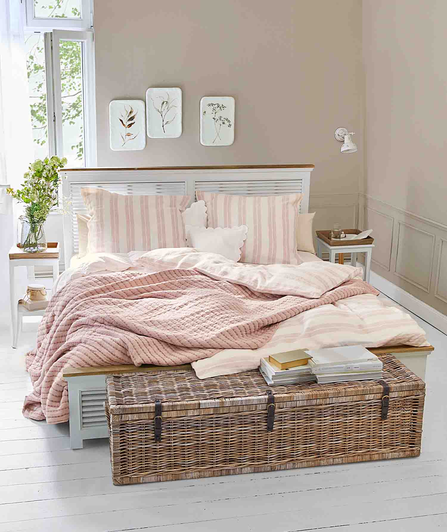 Camera accogliente con letto bianco, coperta trapuntata rosa e baule in vimini con libri sopra