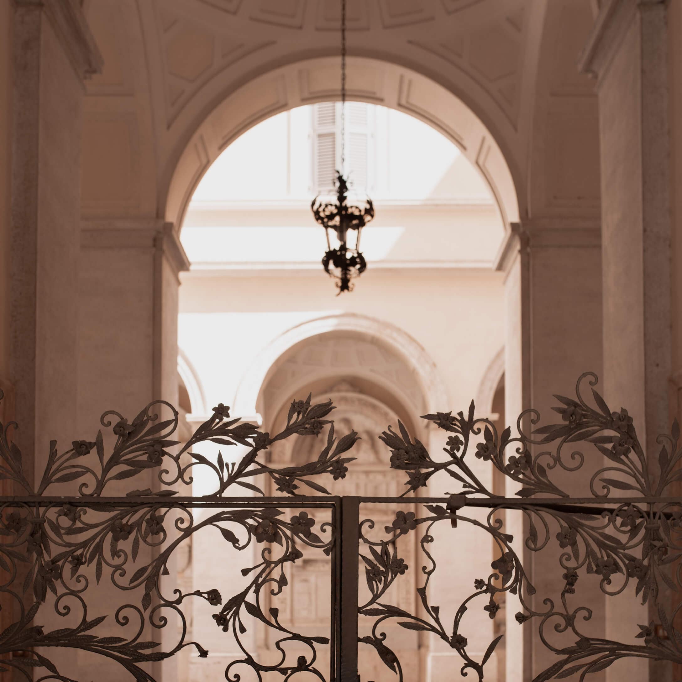 Cancello in ferro battuto decorato con motivi floreali davanti a un arco chiaro con lampada sospesa