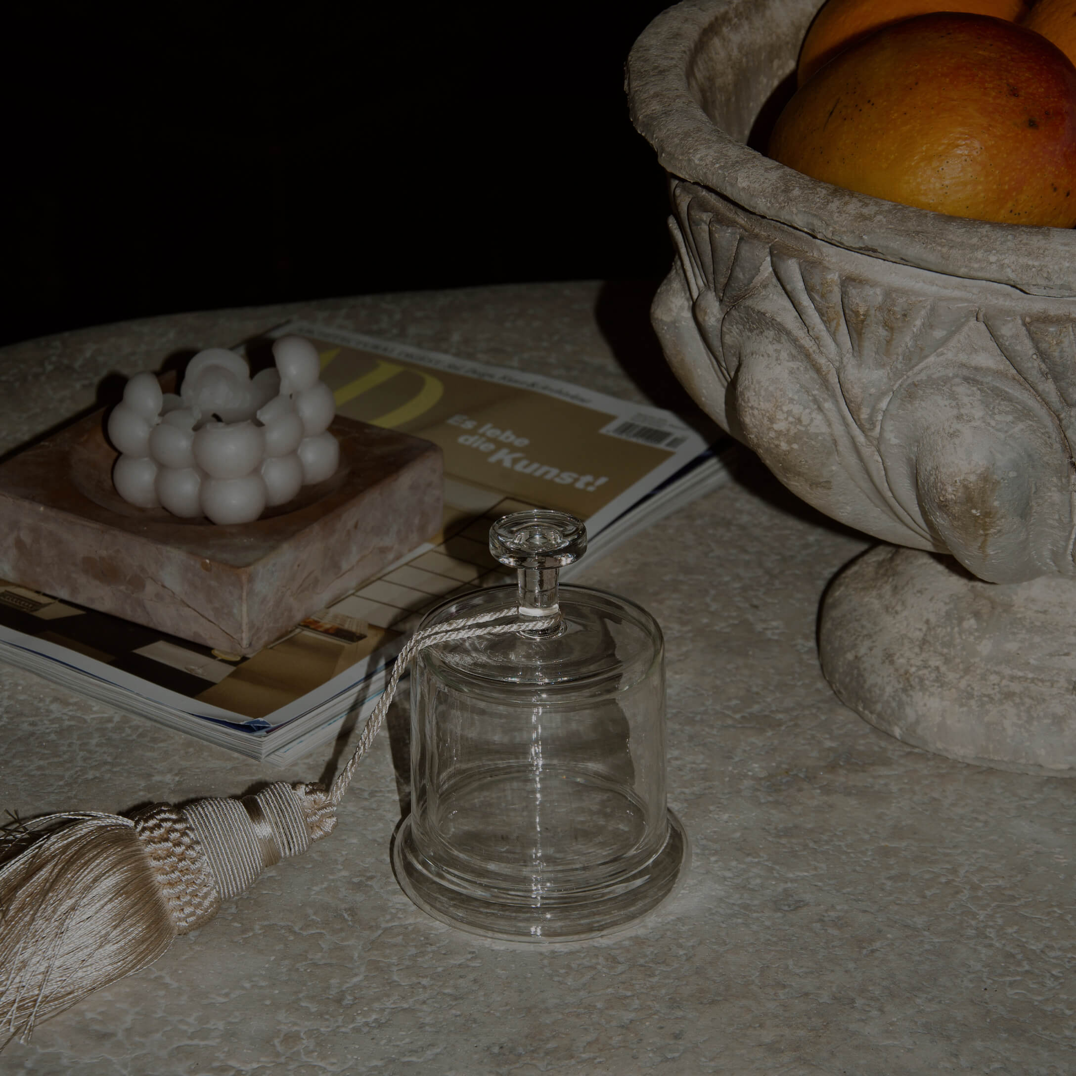 Campana di vetro trasparente con nappina decorativa su tavolo in marmo con candela e ciotola in pietra