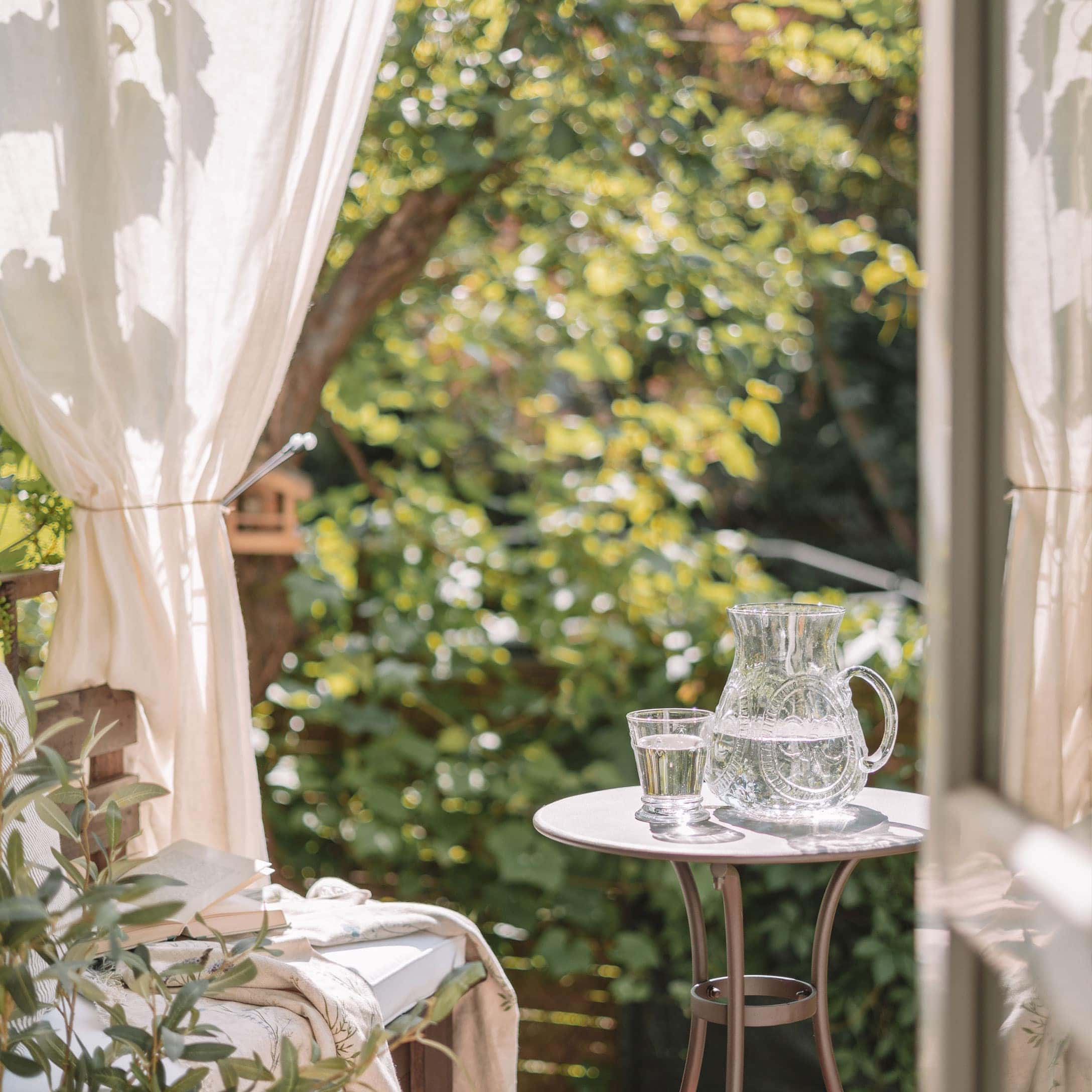 Caraffa e bicchiere di vetro con acqua su tavolo bianco in terrazza con sfondo di piante verdi