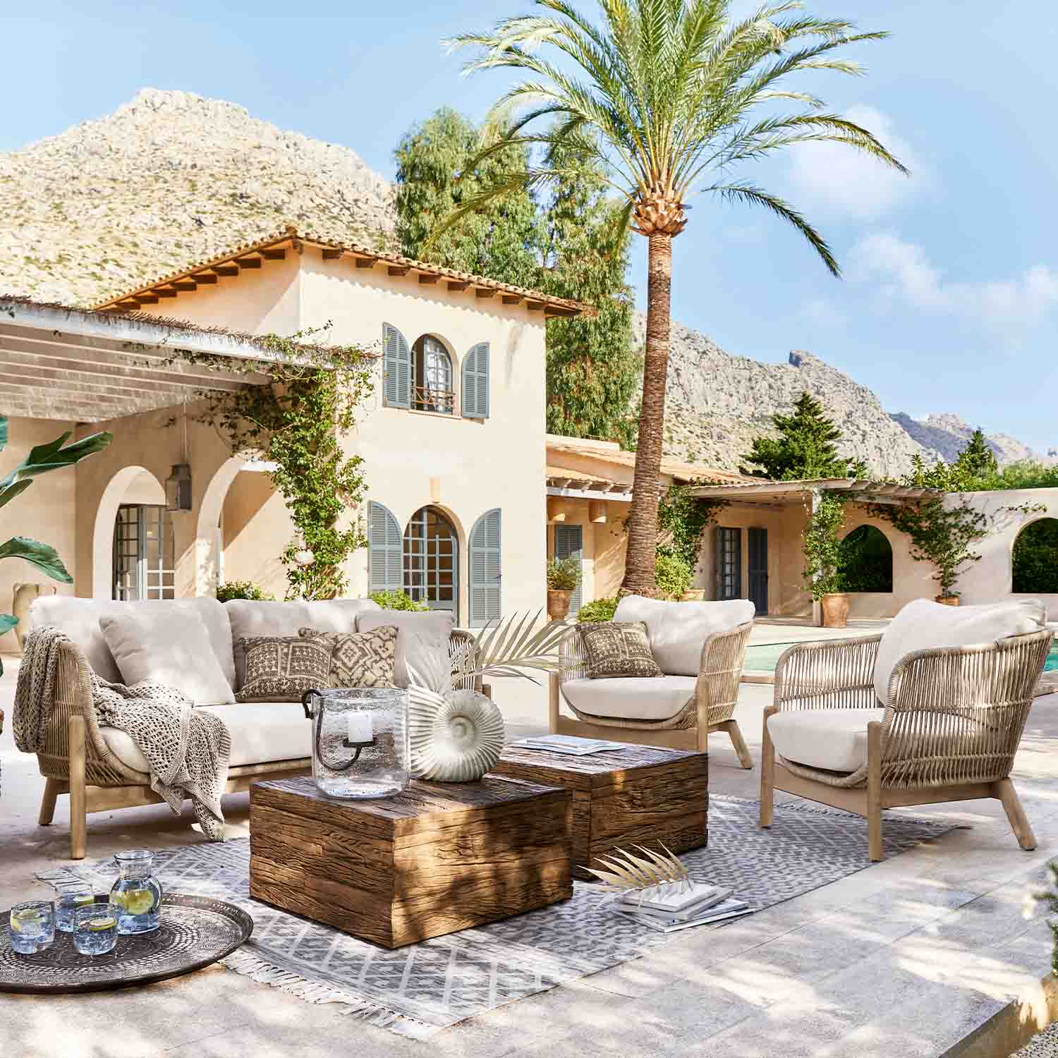 Set di mobili da esterno in rattan chiaro con cuscini beige su terrazza con palma e vista montagna