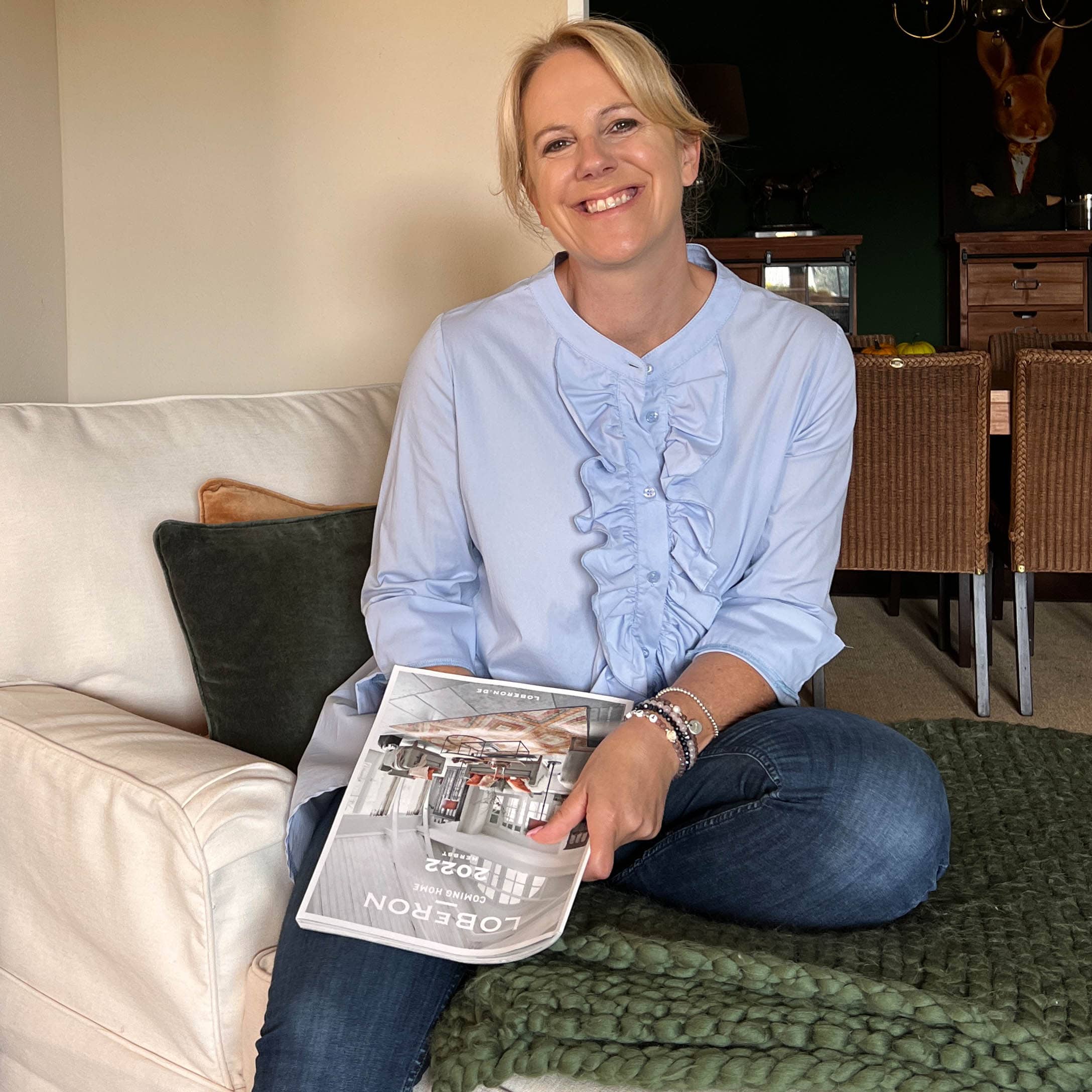 Donna seduta sul divano con rivista Loberon, interno accogliente con coperta lavorata a maglia e cuscini
