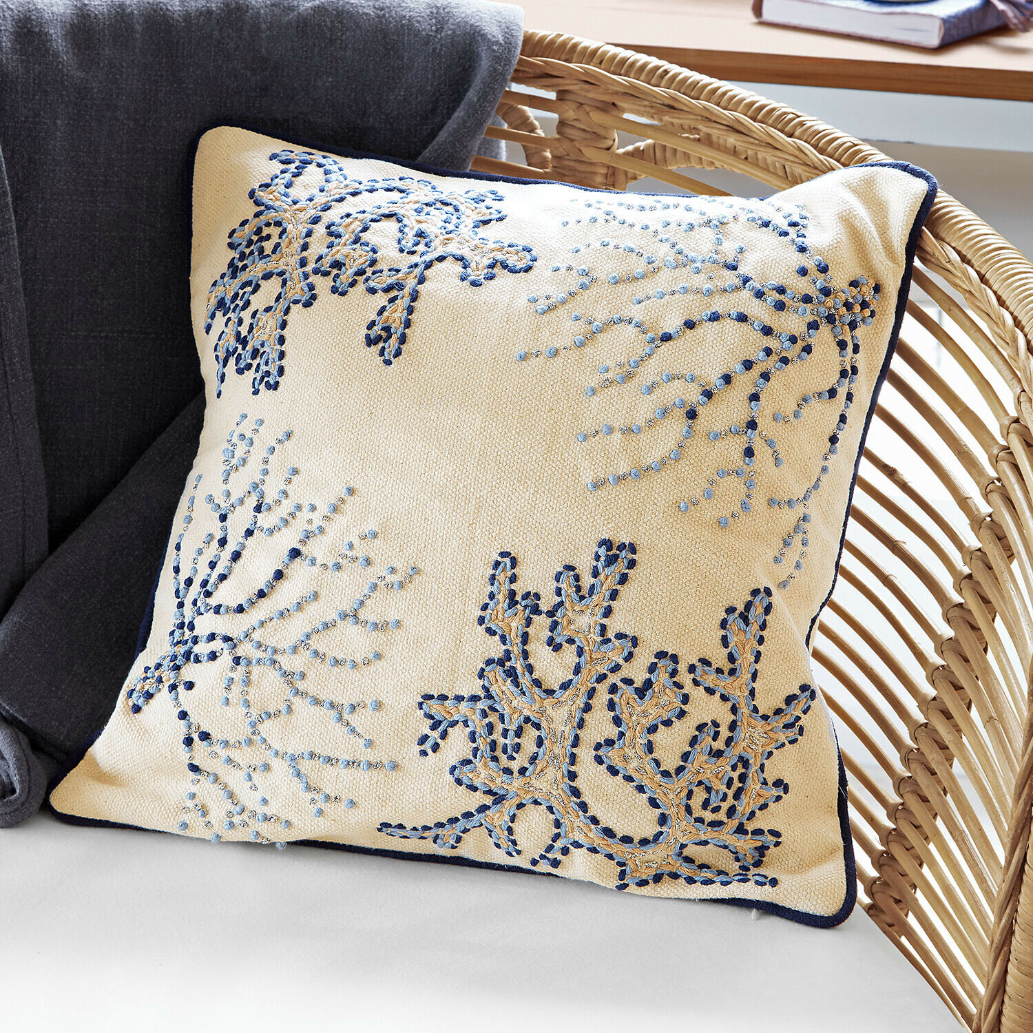 Copricuscino Whitmere Cuscino decorativo beige con ricamo blu e bordo blu scuro su poltrona in vimini