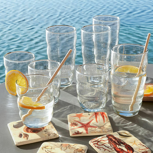Bicchieri in vetro trasparente con fette di limone e mescolatori in legno, sottobicchieri a tema marino vicino all'acqua