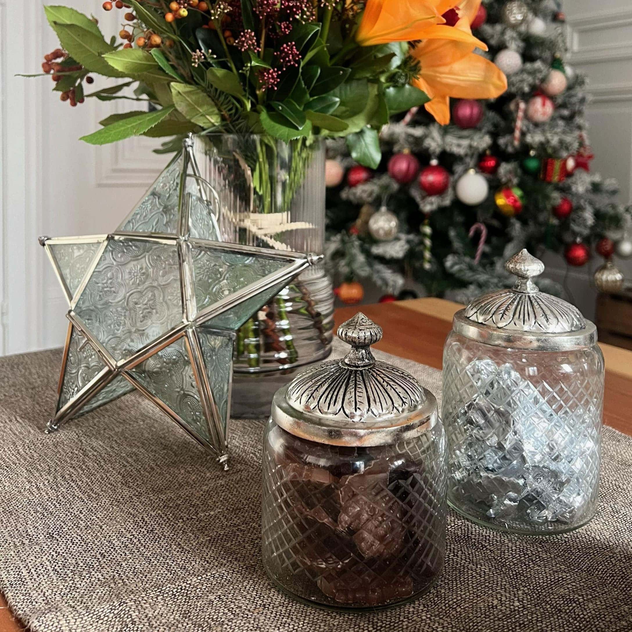 Barattoli di vetro con coperchi in metallo e stella decorativa su tavolo con fiori e albero di Natale sullo sfondo