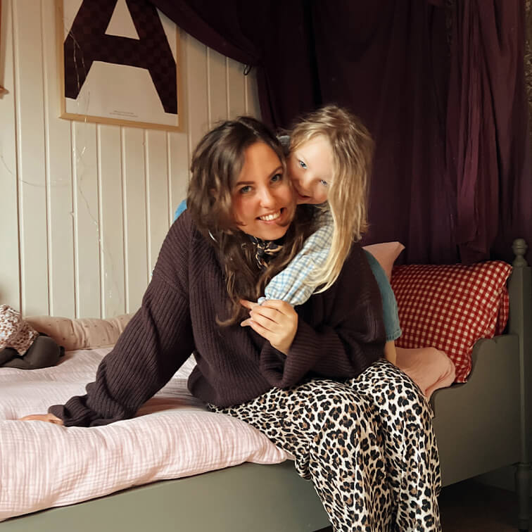 Donna con maglione marrone e pantaloni leopardati seduta sul letto con bambino in camicia chiara a quadri