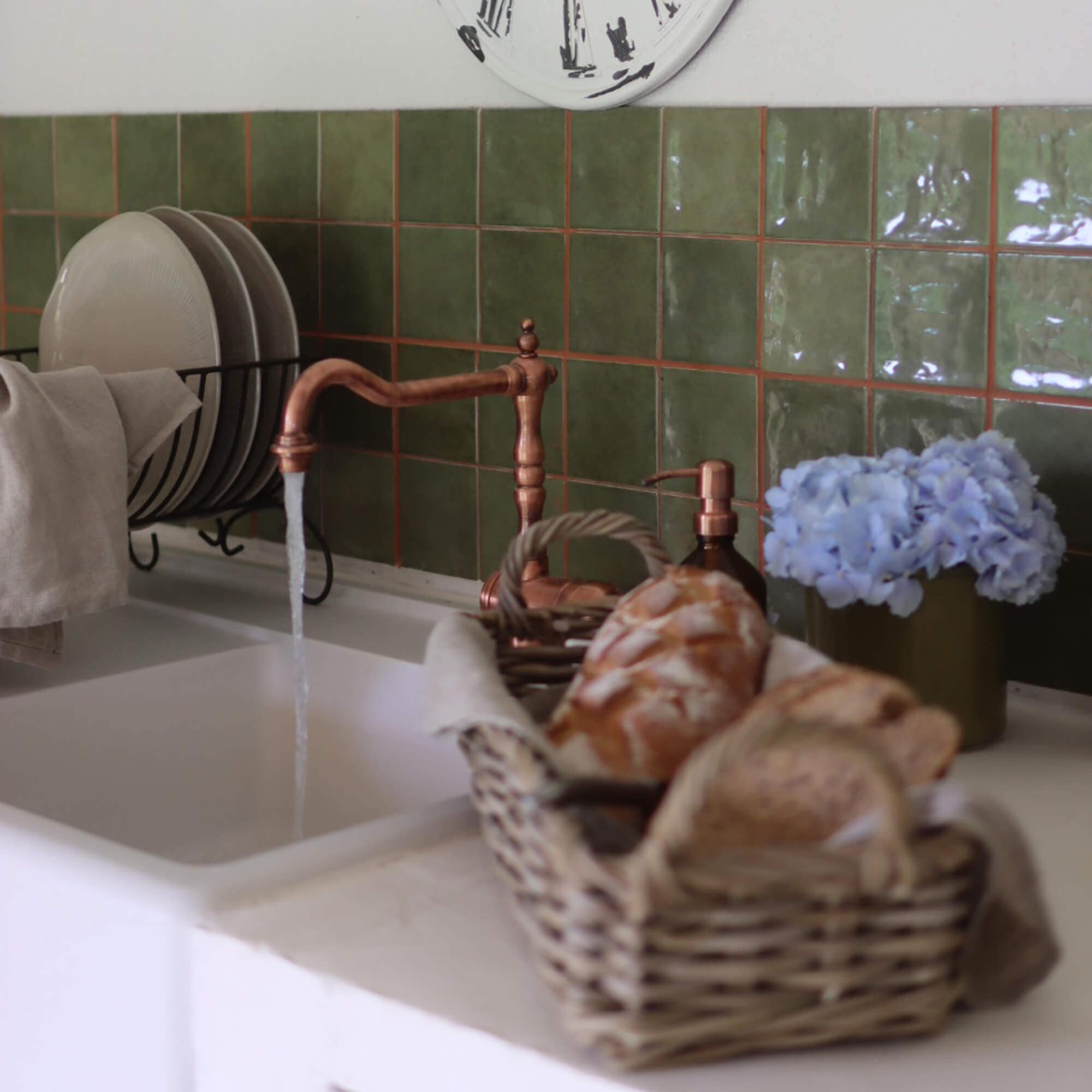 Cucina con rubinetto in rame, piastrelle verdi, cesta di pane e fiori blu decorativi