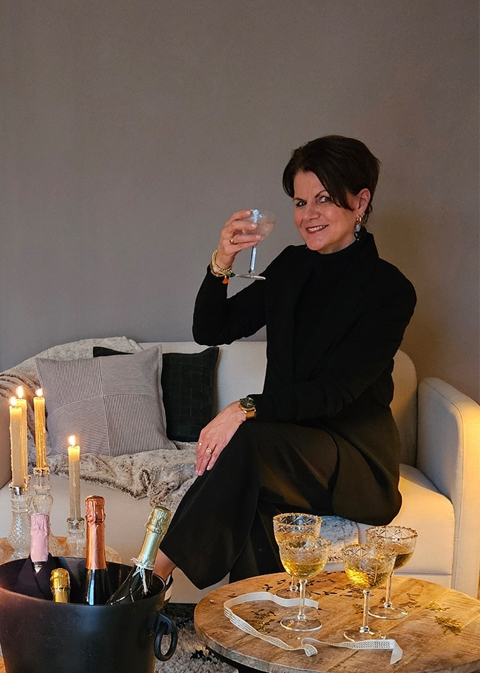 Donna vestita di nero seduta su divano chiaro con bicchiere, candele e bottiglie di champagne accanto