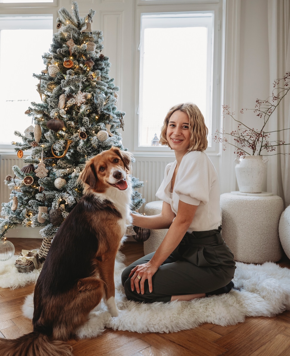 Donna con maglia bianca e pantaloni verdi con cane accanto all'albero di Natale decorato in stanza luminosa