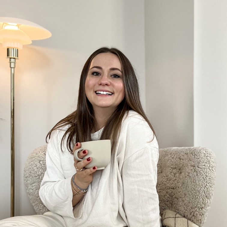 Donna sorridente con maglione bianco che tiene una tazza beige seduta su poltrona morbida in ambiente luminoso