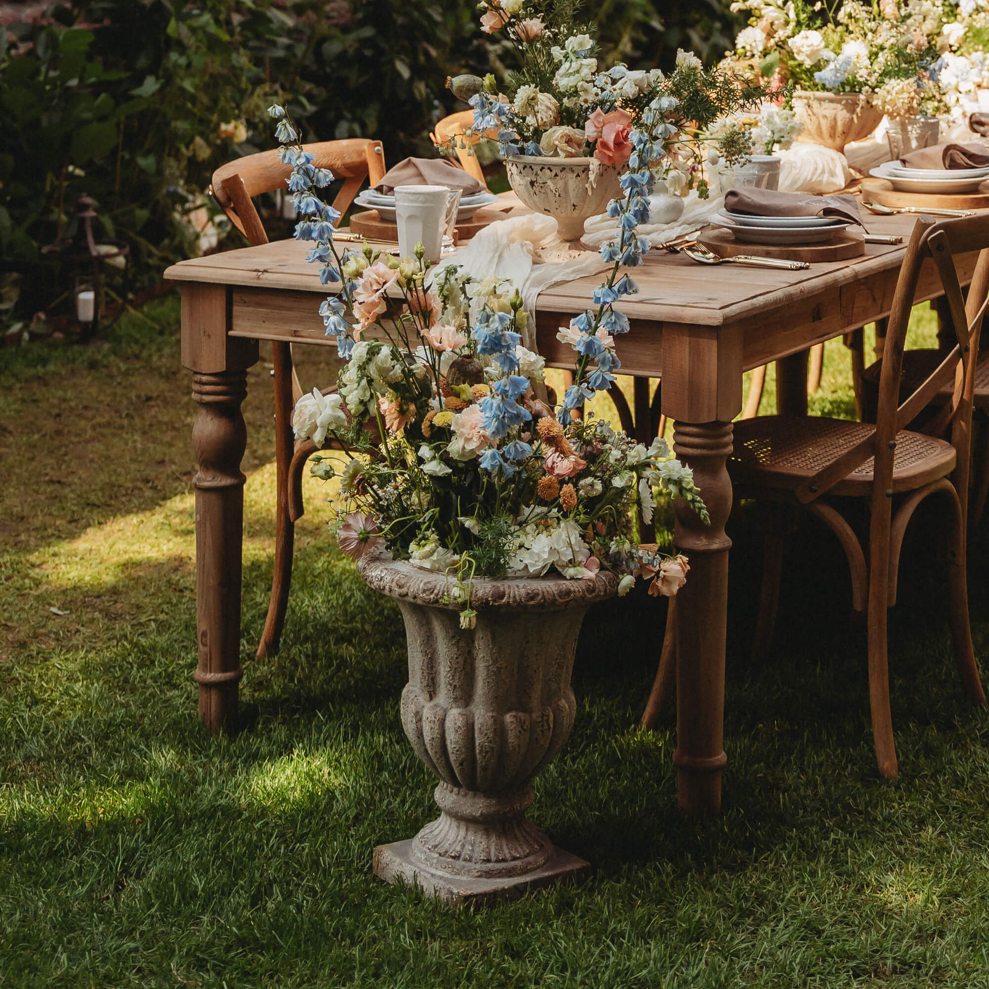 Grande vaso in pietra con fiori pastello davanti a tavolo e sedie in legno in giardino