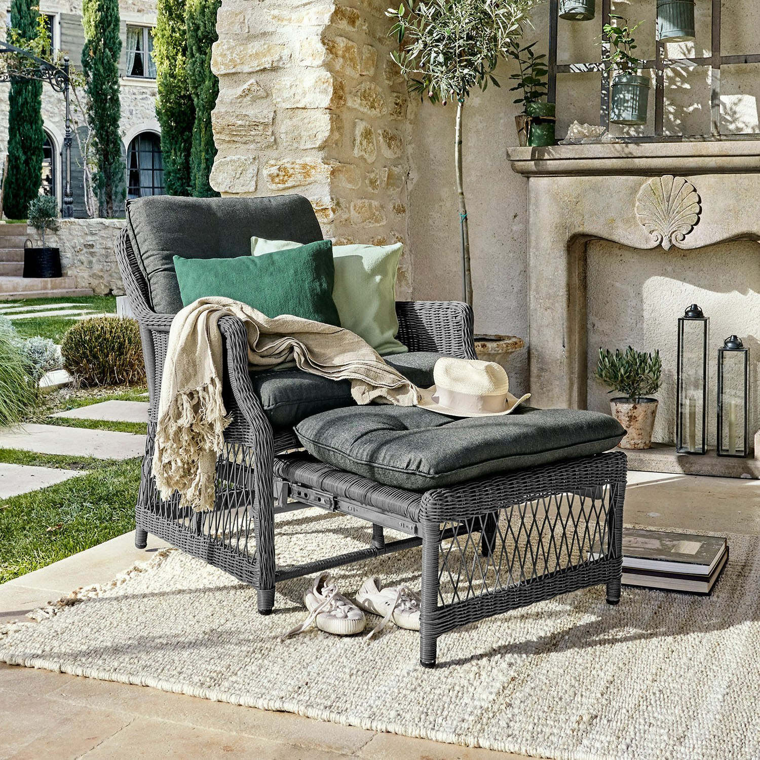 Poltrona Howe Poltrona in rattan grigio con poggiapiedi e cuscini grigio scuro, decorata con cuscini verdi su una terrazza