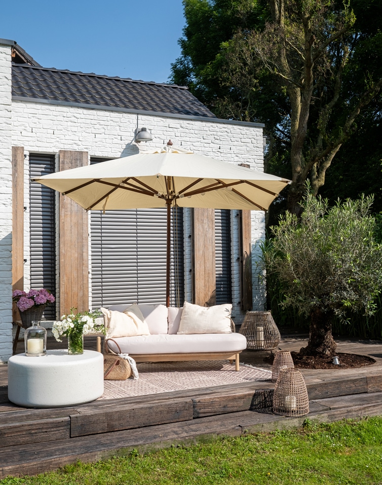 Terrazza con muro in mattoni bianchi, divano beige con cuscini e ombrellone crema su terrazza in legno