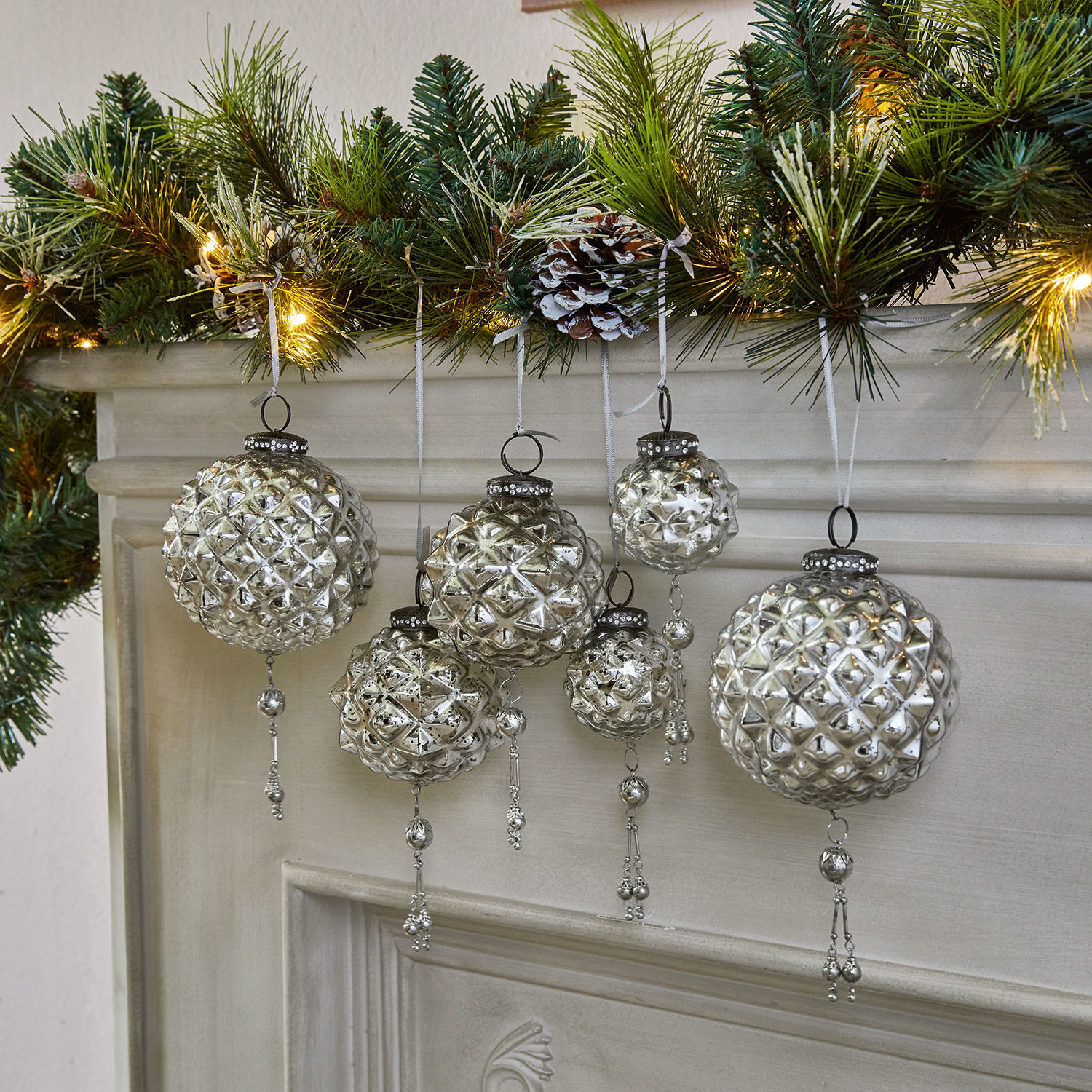 Set di 6 decorazioni natalizie Murron Palle di Natale in vetro argentato con decorazioni appese davanti a un camino e rami verdi con luci