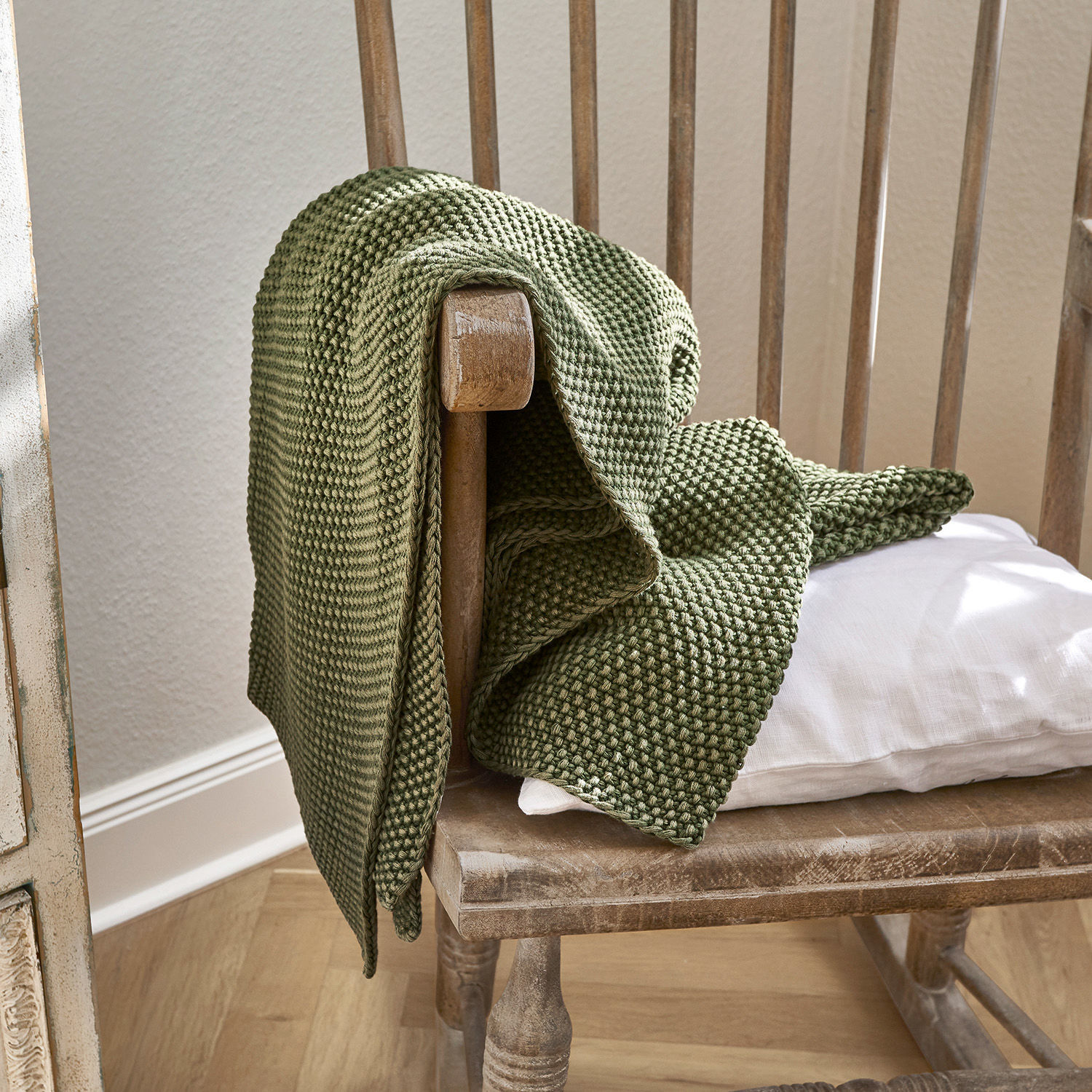 Plaid Amberley Coperta verde in cotone lavorata a maglia su sedia in legno con cuscino bianco, decorazione accogliente