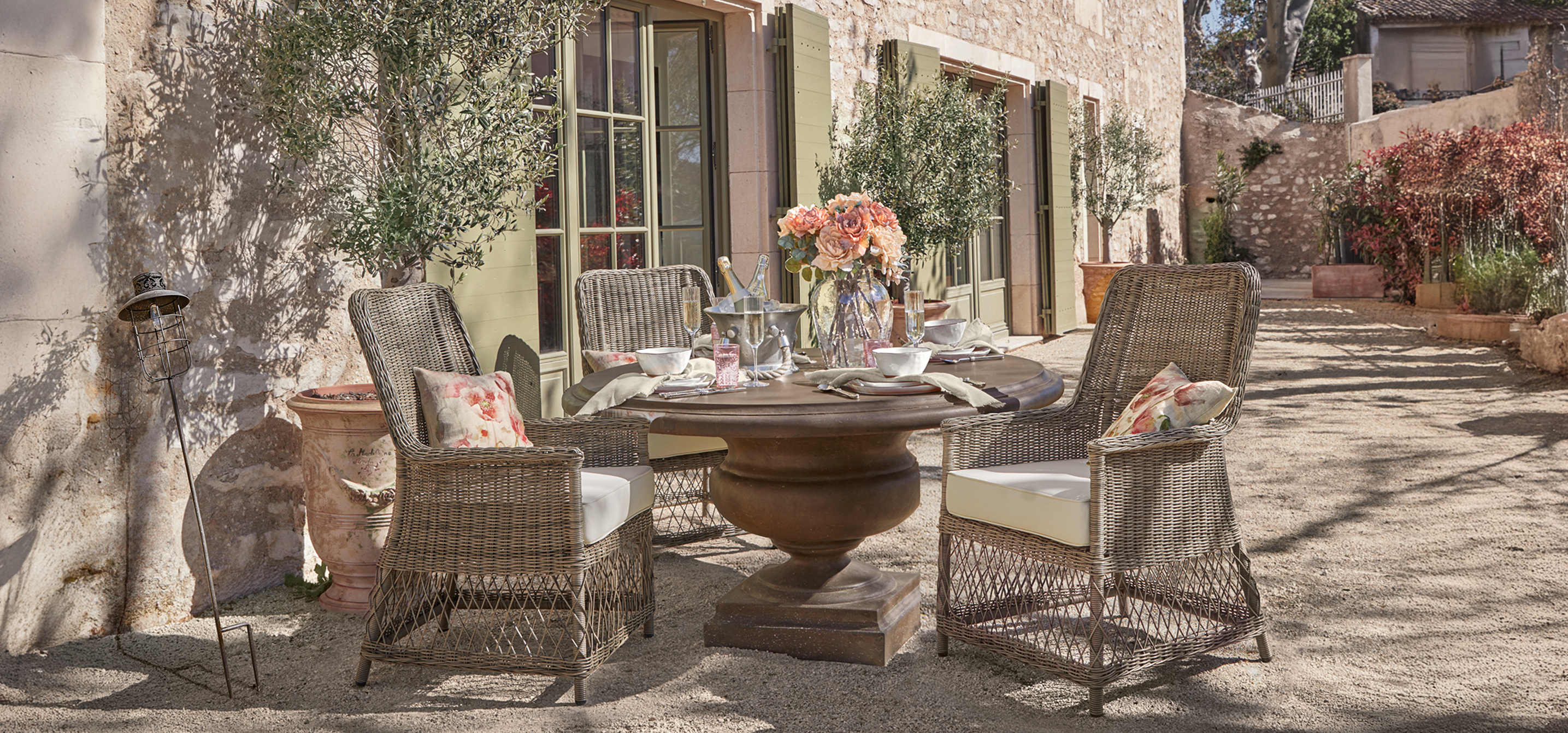 Tavolo da giardino rotondo in metallo marrone con quattro sedie in vimini e cuscini beige in terrazza soleggiata