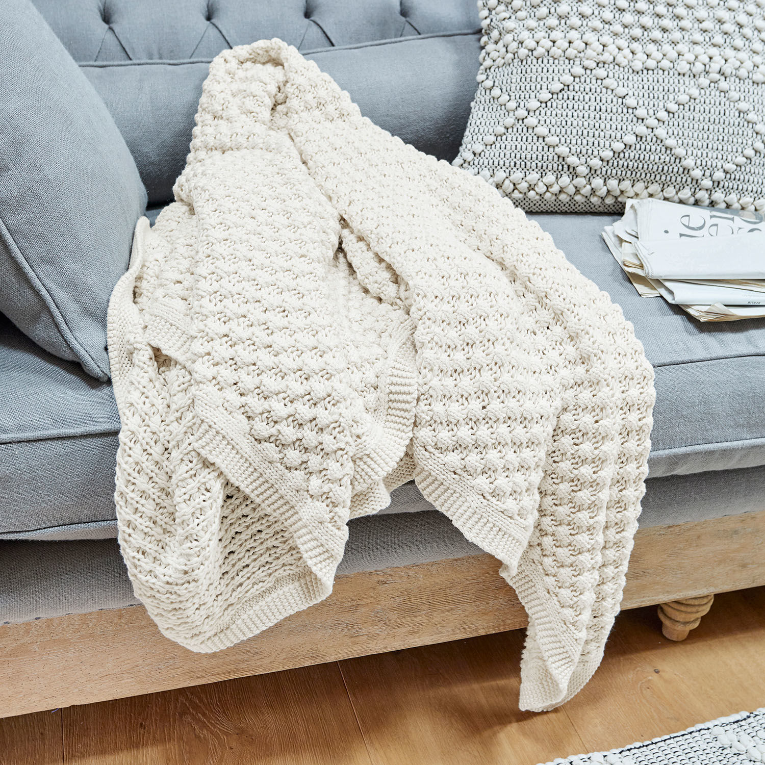 Coperta Tremes Coperta in cotone lavorata a maglia color crema su divano grigio con cuscino decorativo e giornali
