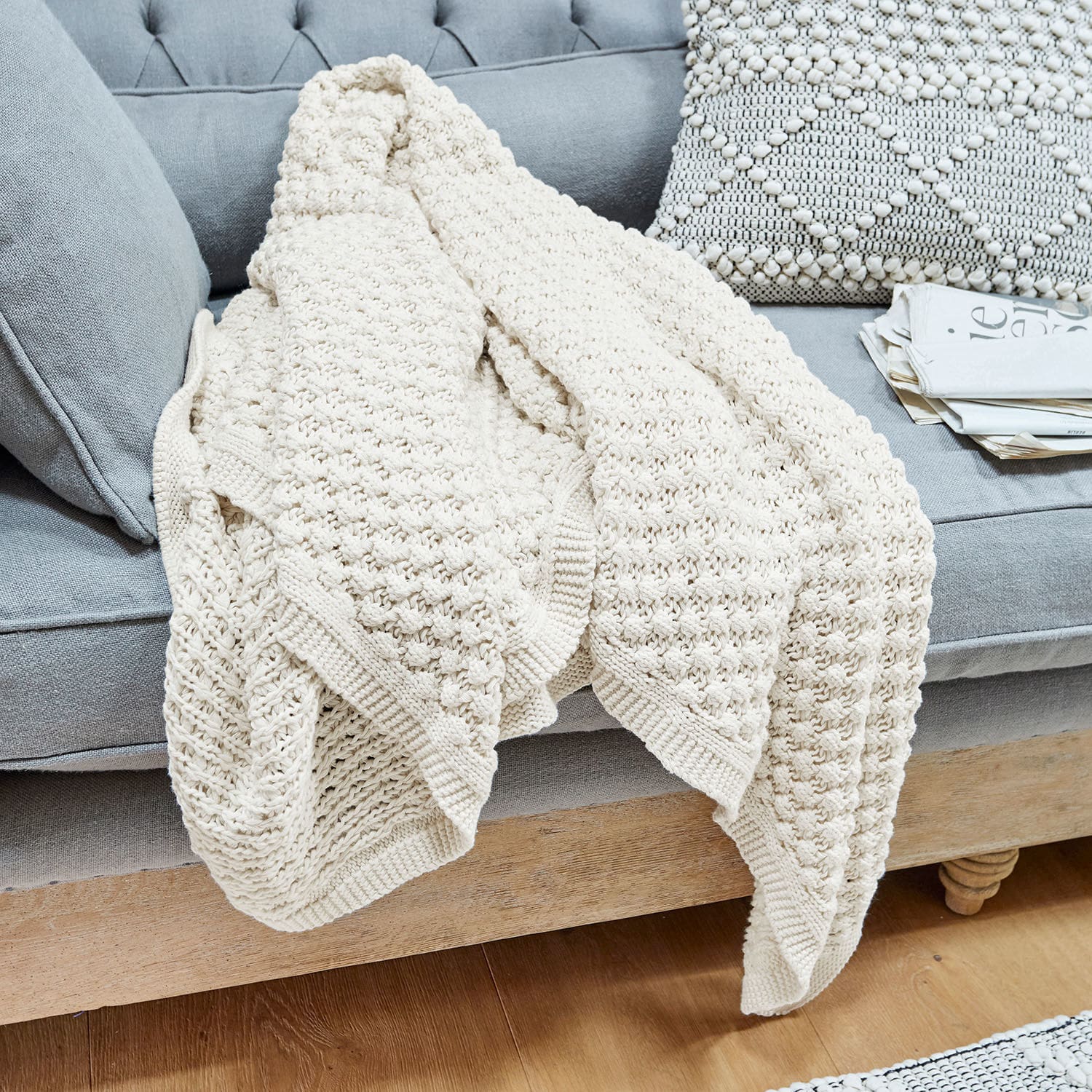 Coperta Tremes Coperta in cotone lavorata a maglia color crema su divano grigio con cuscino decorativo e giornali