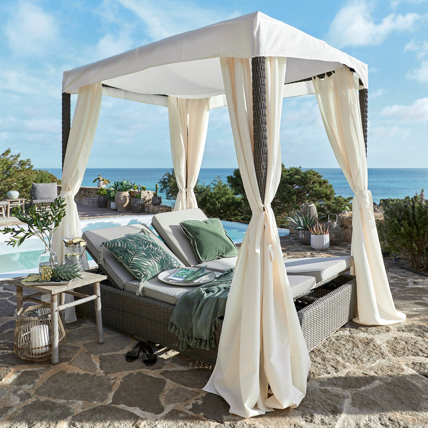 Lounge da giardino Hereford Lettino da giardino in rattan con tenda crema su terrazza in pietra con vista mare