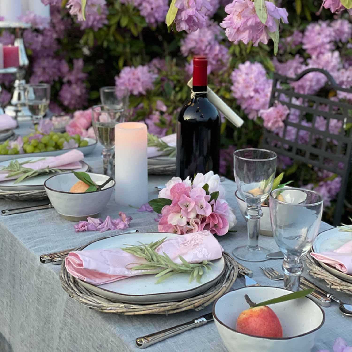 Tavola elegantemente apparecchiata con stoviglie bianche, tovaglioli rosa e bottiglia di vino rosso in giardino fiorito