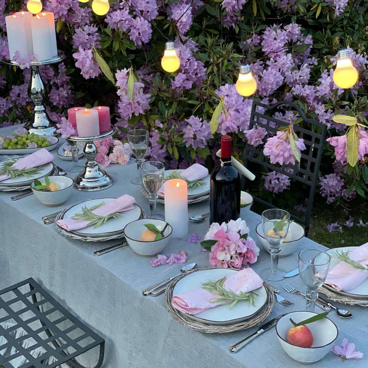 Tavolo da giardino elegantemente apparecchiato con tovaglioli rosa, candele e bottiglia di vino davanti a fiori viola