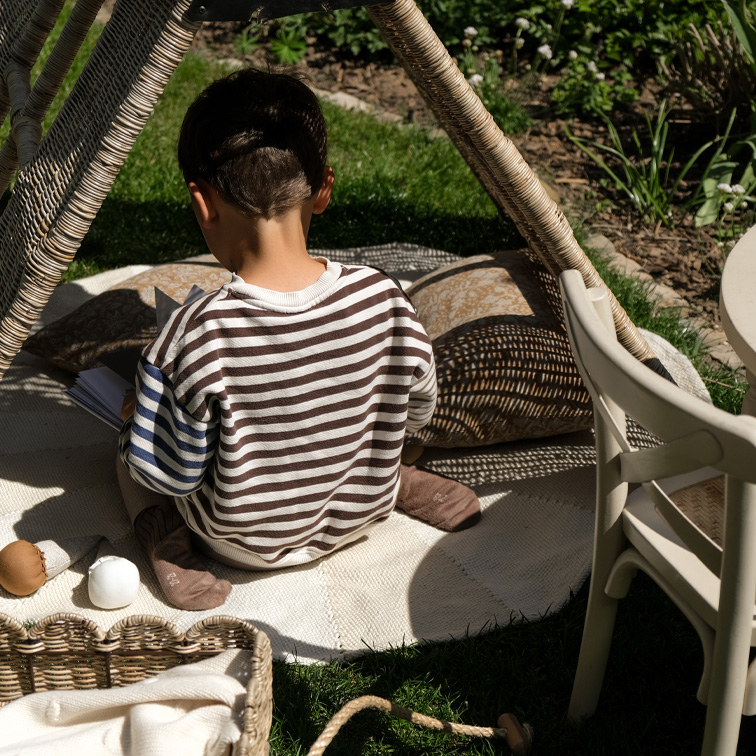 Bambino con maglione a righe seduto su tappeto sotto tenda in vimini in giardino, momento di relax