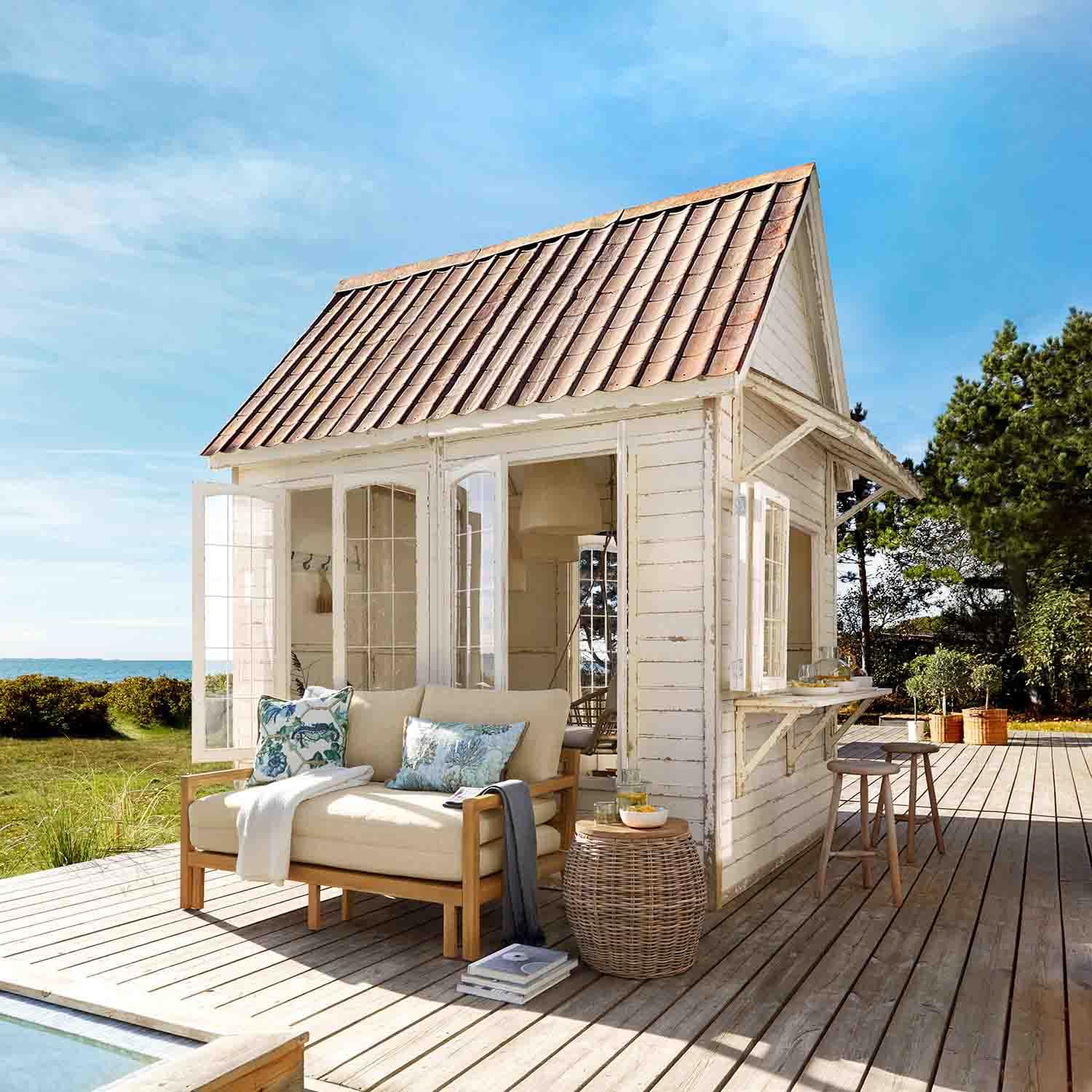 Casetta in legno bianco con tetto rosso su terrazza in legno con divano beige e tavolo in vimini
