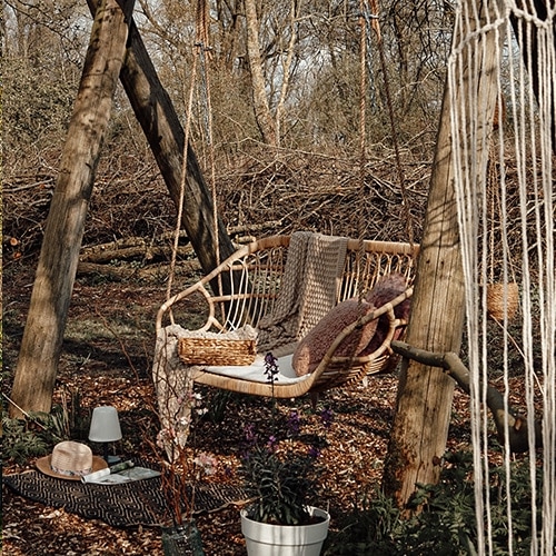 Altalena da giardino in rattan naturale con cuscini e coperta su sfondo boschivo autunnale