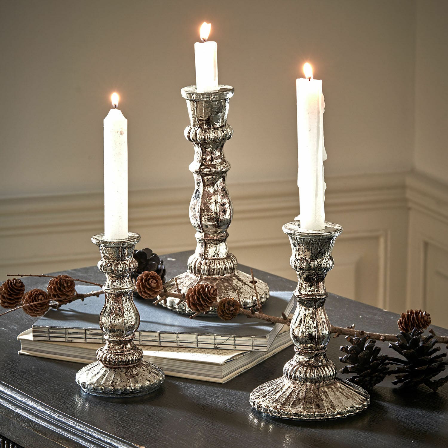 Set di 3 candelieri Favalina Tre candelabri vintage in metallo argento con candele bianche su tavolo scuro decorato con pigne