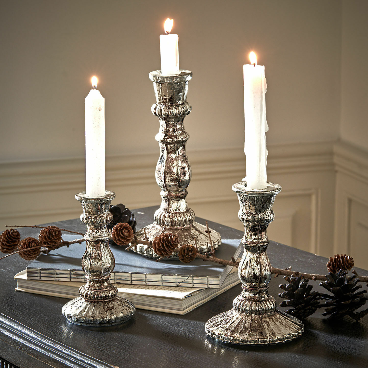 Set di 3 candelieri Favalina Tre candelabri vintage in metallo argento con candele bianche su tavolo scuro decorato con pigne