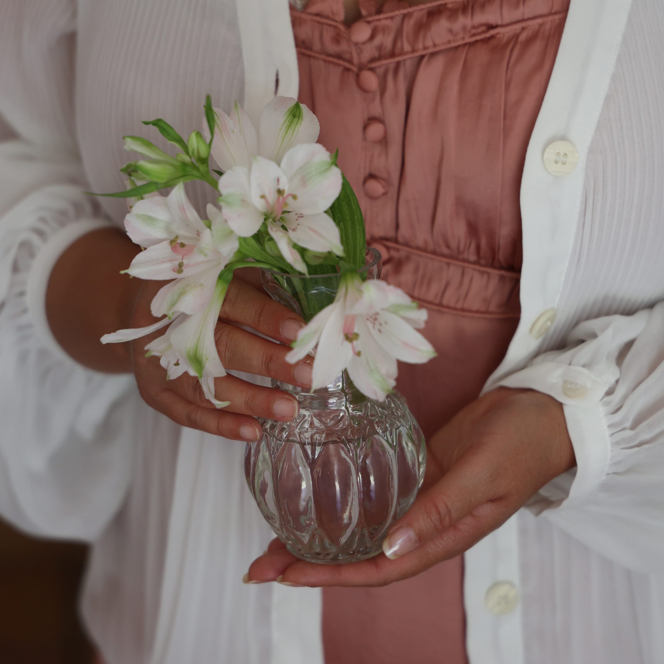 Donna che tiene un vaso di vetro trasparente con fiori bianchi davanti a un vestito rosa e cardigan bianco