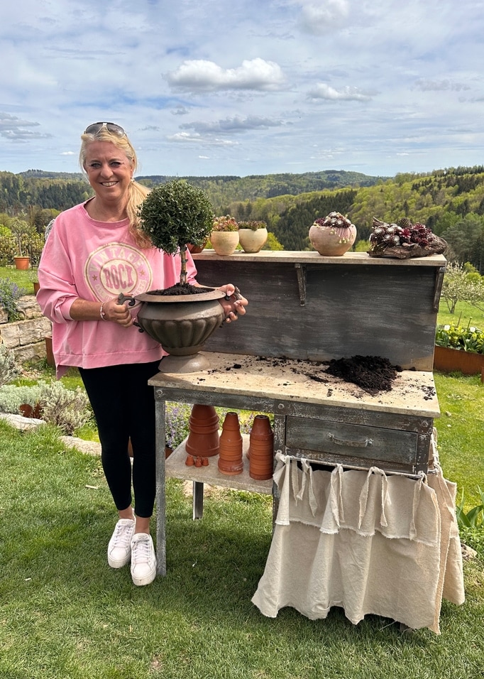 Donna con felpa rosa tiene vaso con cespuglio verde vicino a tavolo da giardino in legno con paesaggio verde