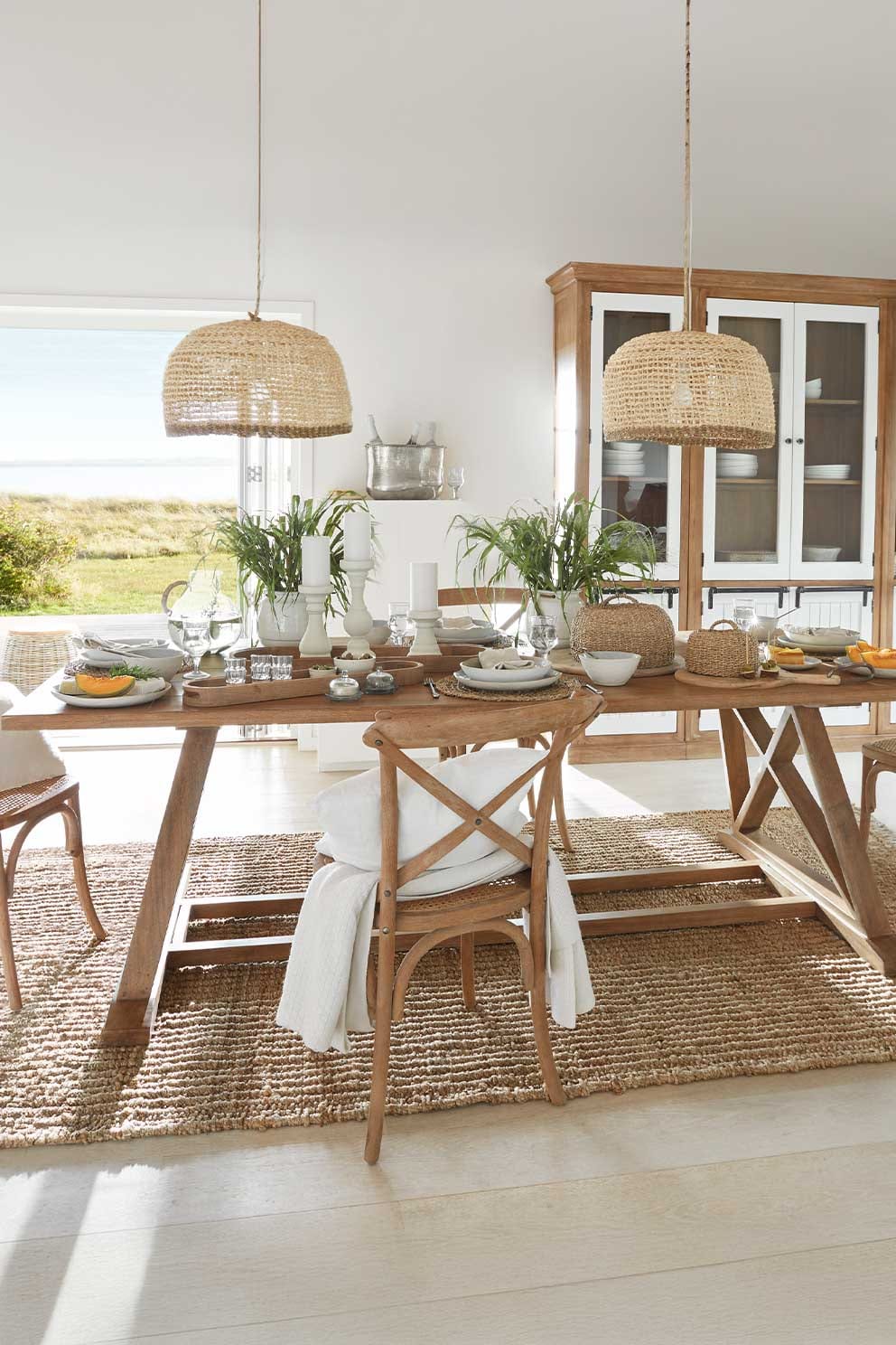 Sala da pranzo con tavolo e sedie in legno, lampade a sospensione in rattan naturale, piante decorative e accessori bianchi