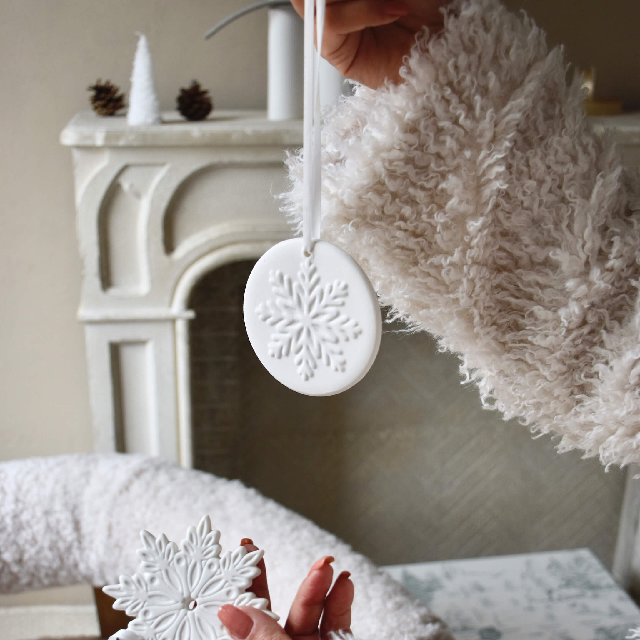 Decorazioni natalizie in ceramica bianca con motivo fiocco di neve, tenute davanti a un camino e maglione morbido