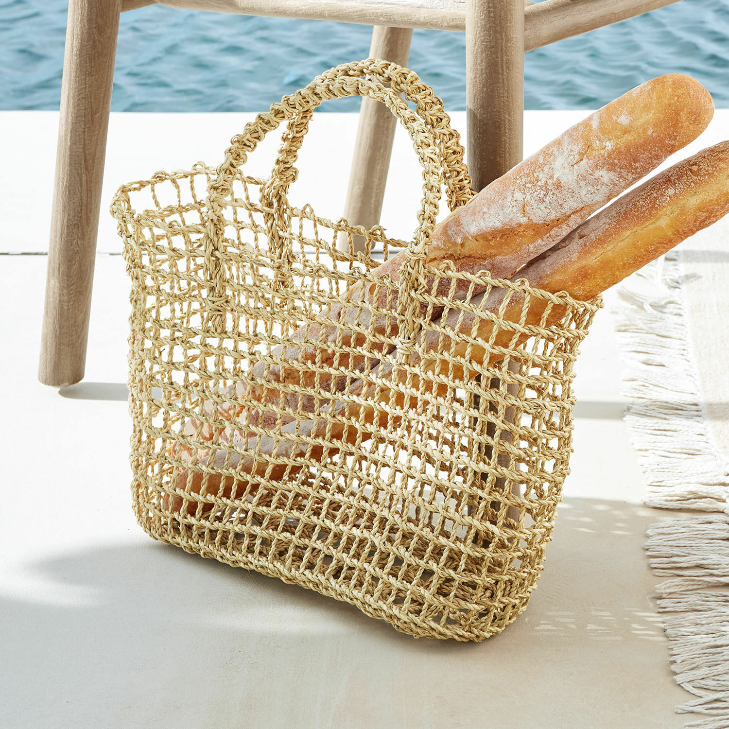 Borsa Xaintieux Borsa rete naturale in materiale intrecciato con due baguette accanto a uno sgabello in legno