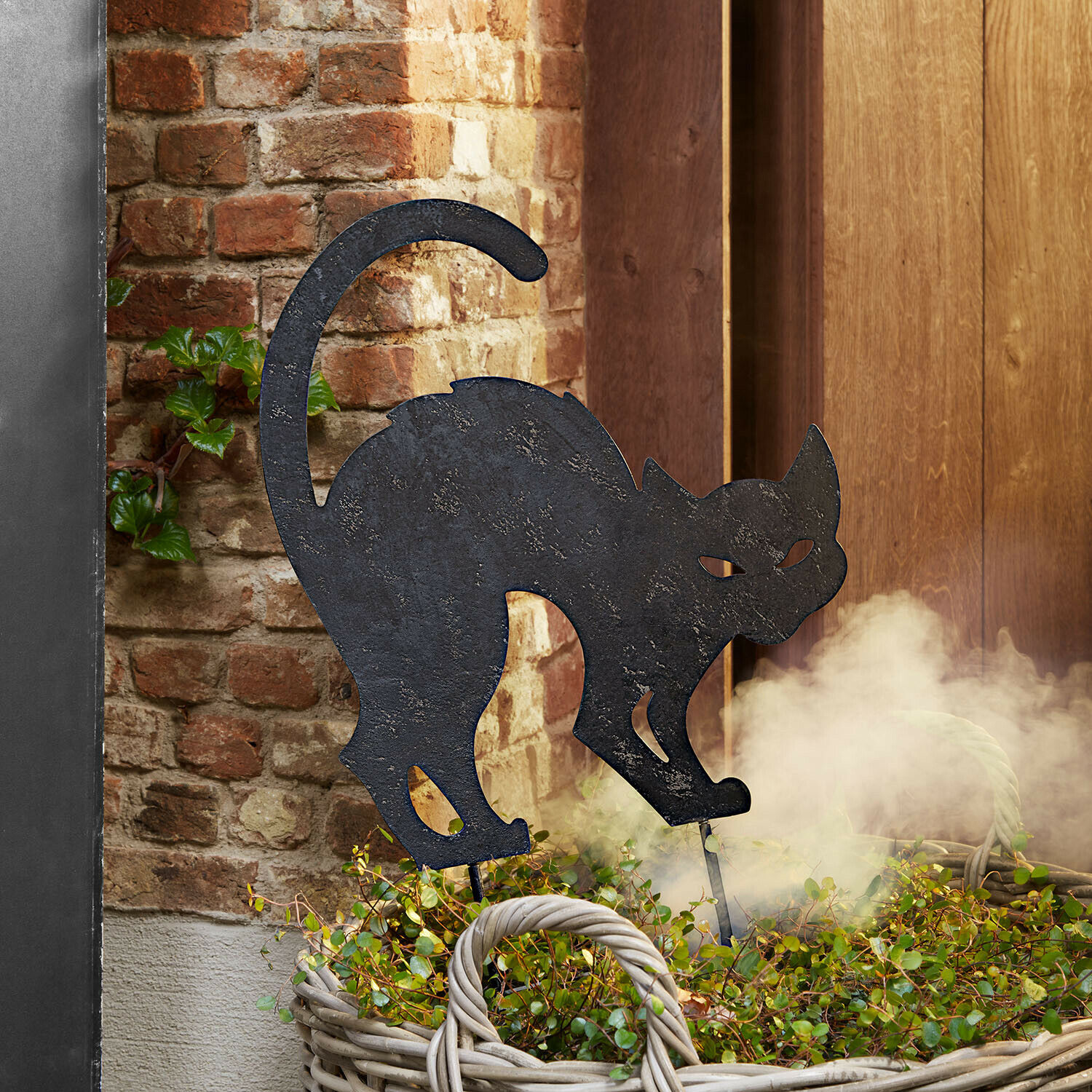 Asta da giardino Rénelise Sagoma decorativa di gatto in metallo nero in un cesto con piante verdi davanti a muro di mattoni