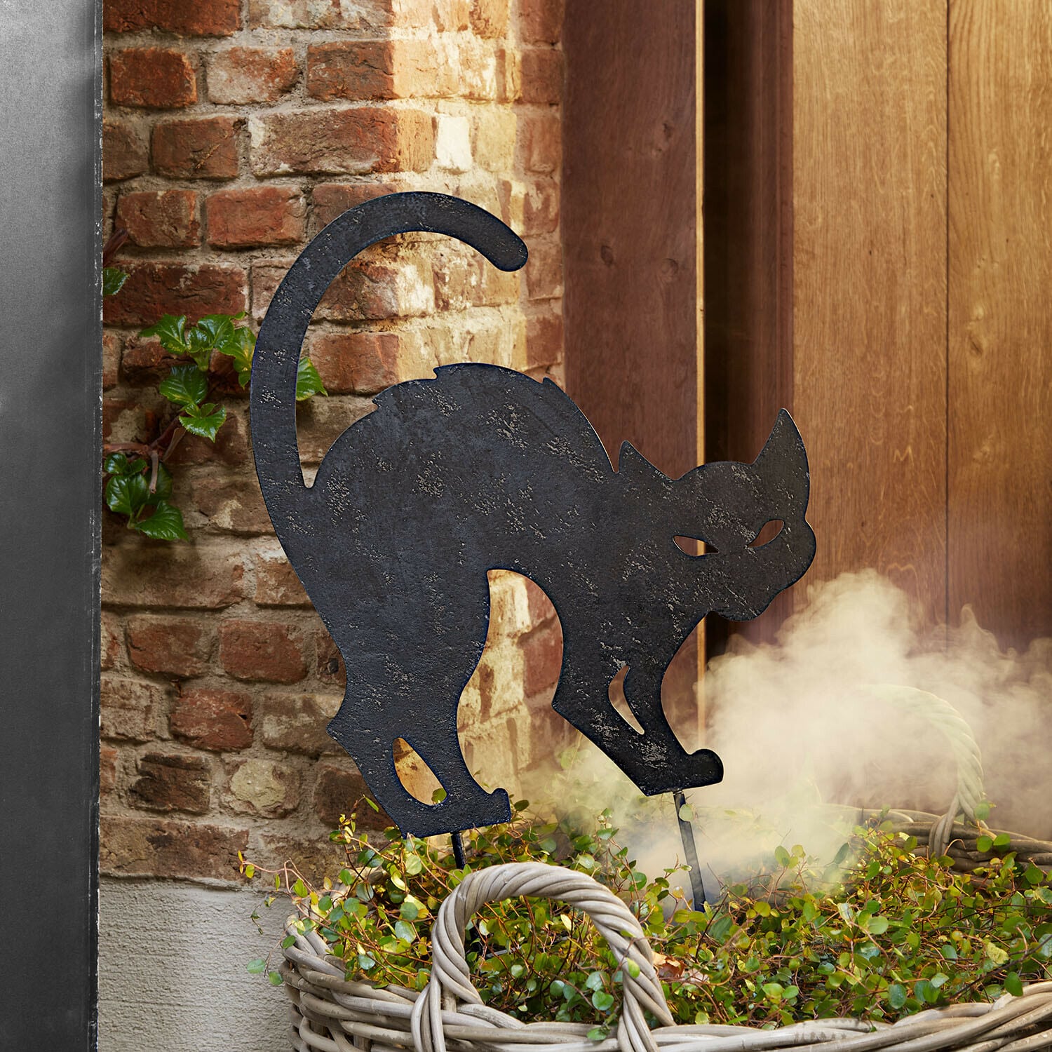 Asta da giardino Rénelise Sagoma decorativa di gatto in metallo nero in un cesto con piante verdi davanti a muro di mattoni