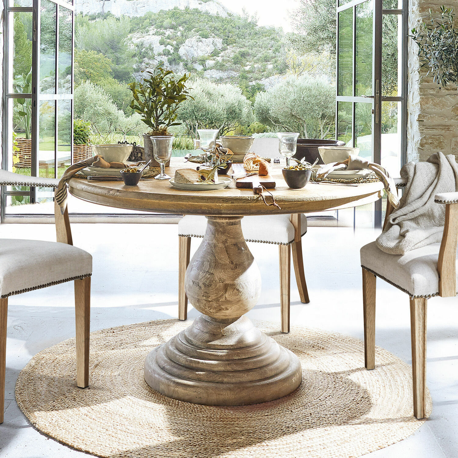 Tavolo Orlin Tavolo rotondo in legno con base decorativa su tappeto naturale in sala da pranzo luminosa con vista sul giardino