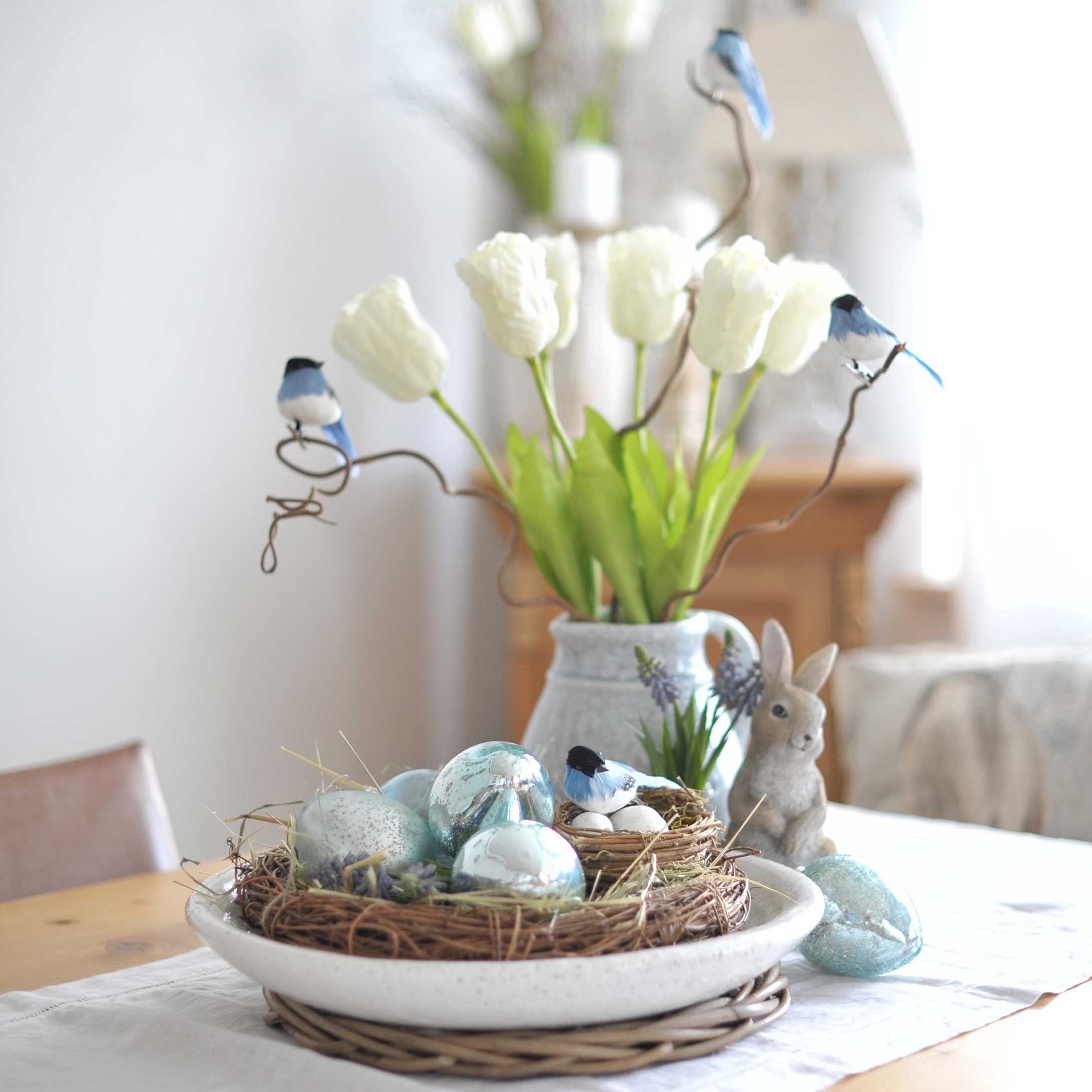 Corona decorativa con uova azzurre e uccellini su tavolo con tulipani bianchi e figura di coniglio