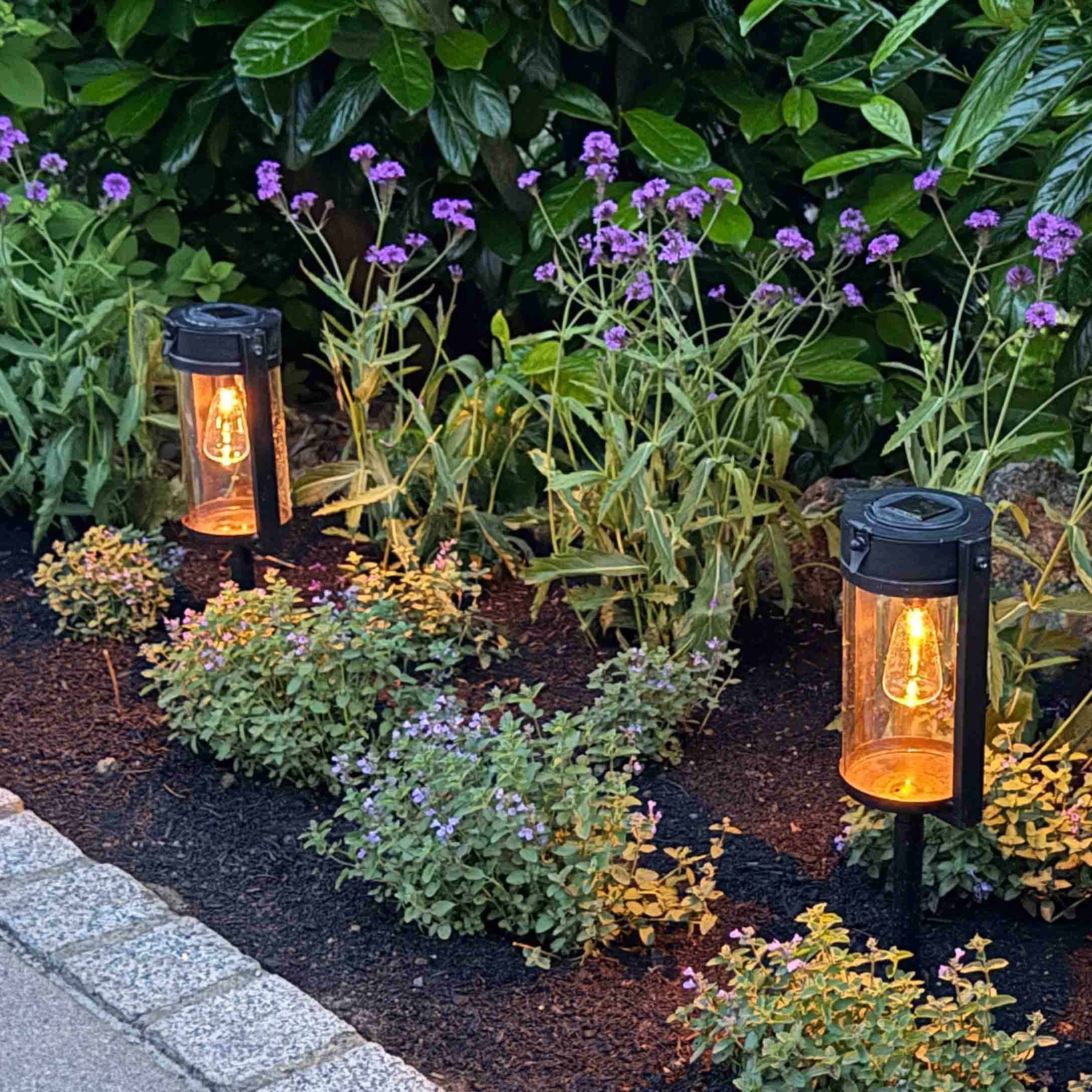 Illuminazione da giardino in metallo nero con luce calda tra piante verdi e fiori viola