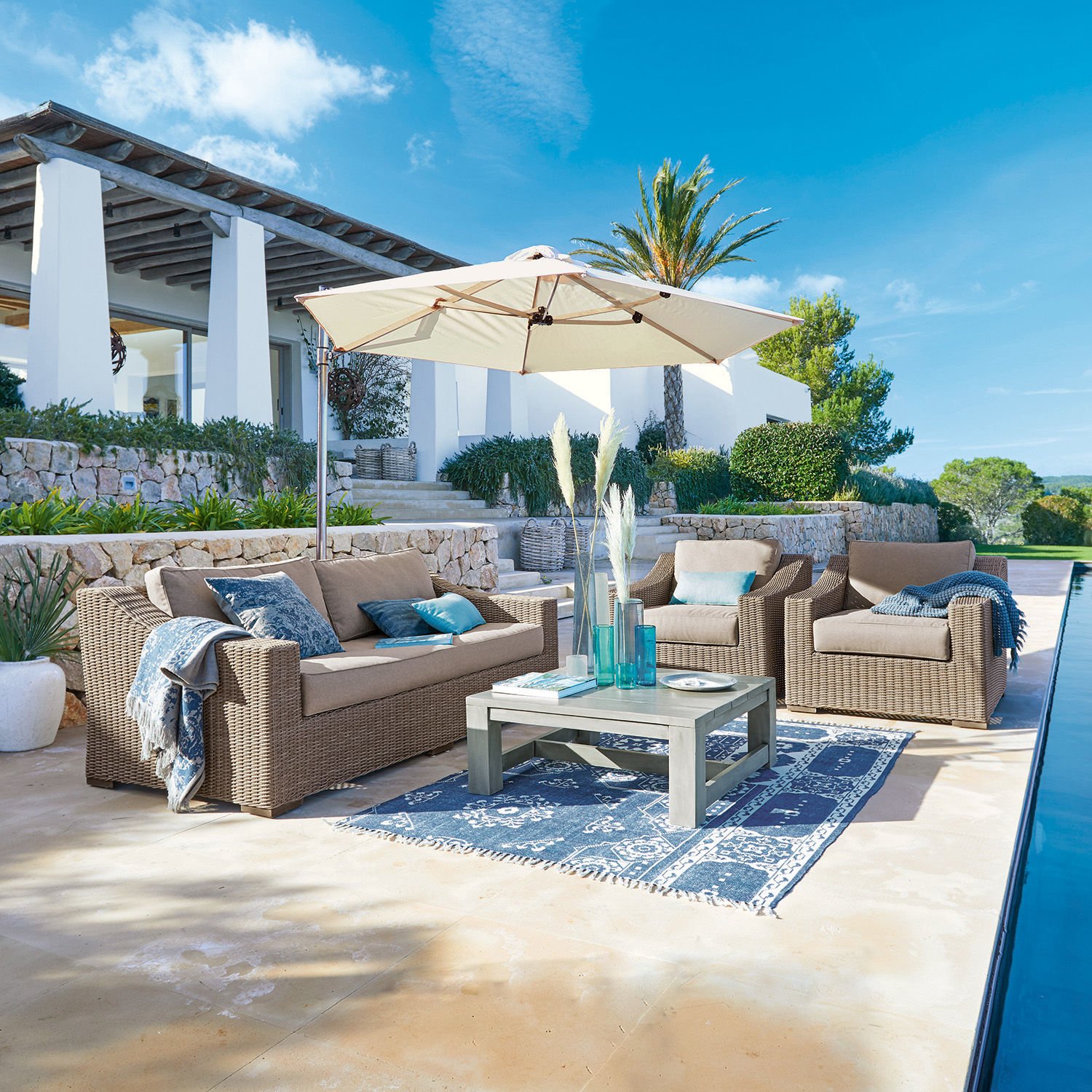 Salottino Chestingdale Set di mobili da giardino in rattan beige con ombrellone e tappeto blu su terrazza soleggiata
