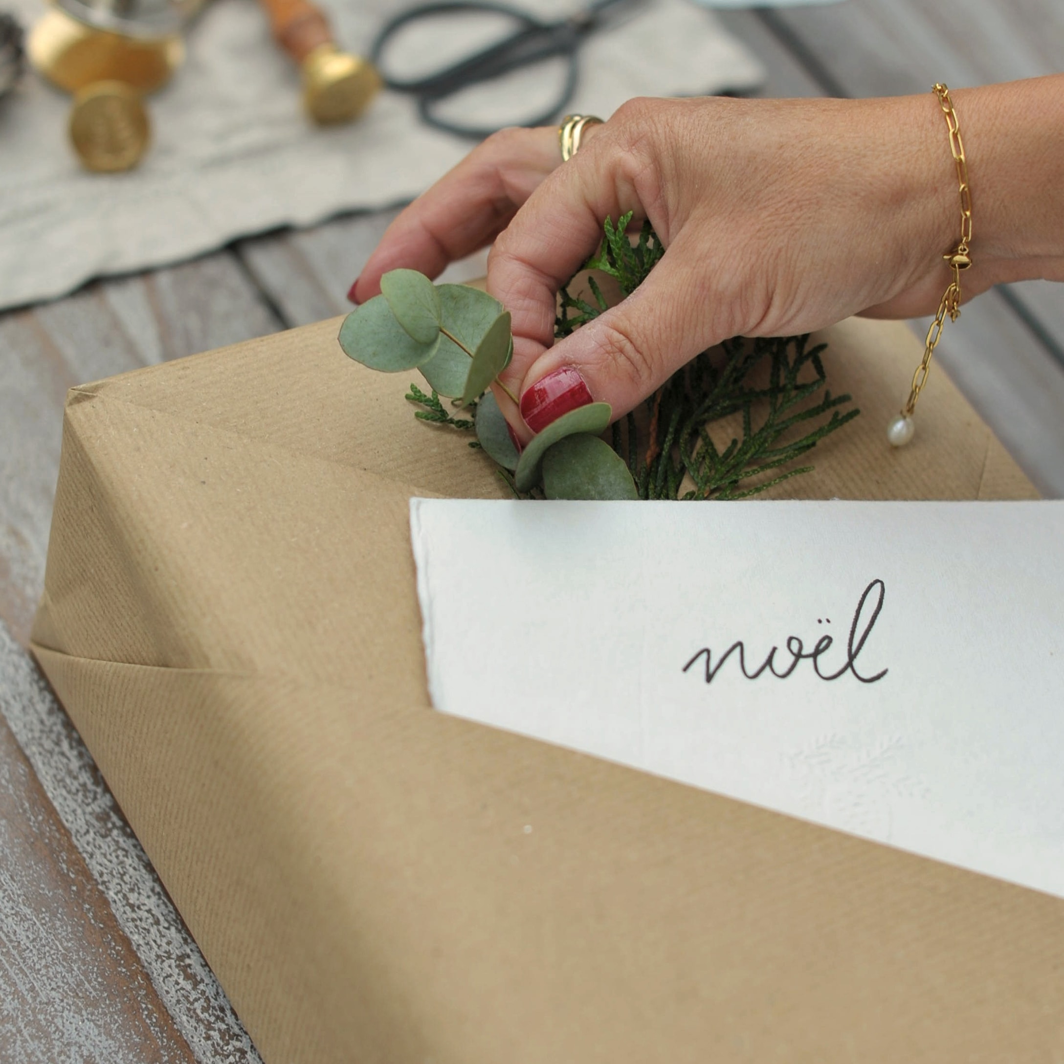 Mano che decora regalo con carta kraft marrone, foglie verdi e biglietto scritto Noël