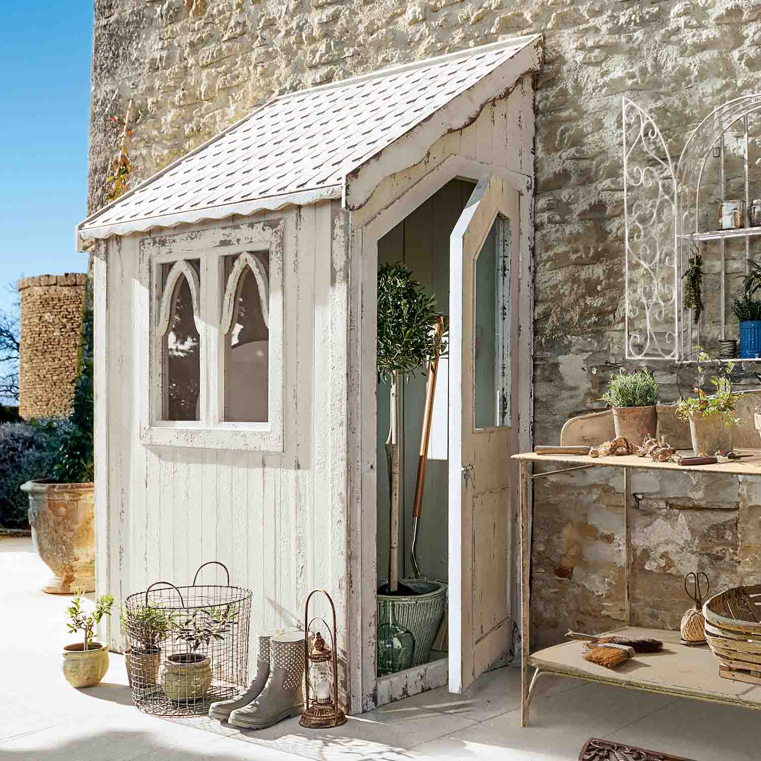 Piccolo capanno da giardino in legno bianco con finestre e porta aperta, attrezzi e decorazioni