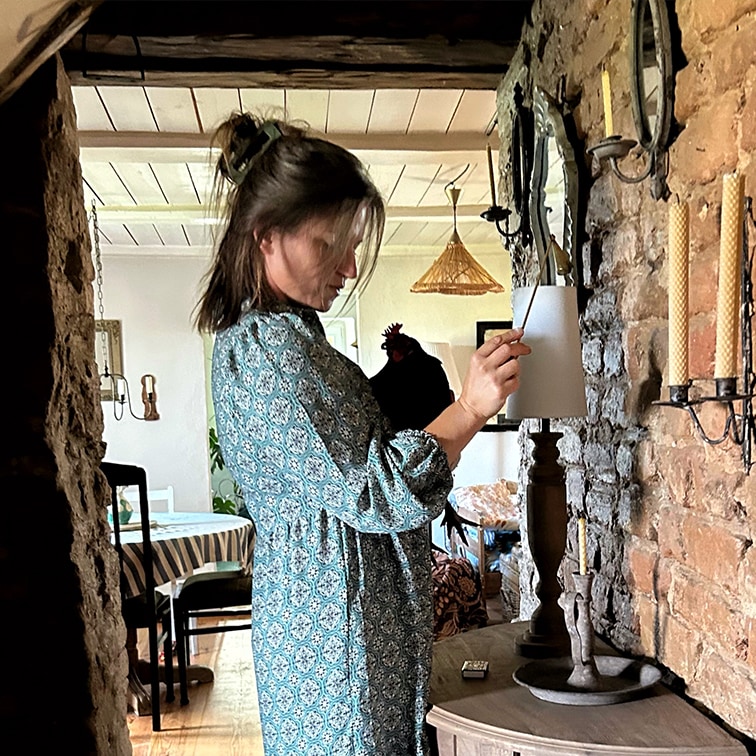 Donna con vestito blu a motivi accende lampada in interno rustico con muro in mattoni e candele