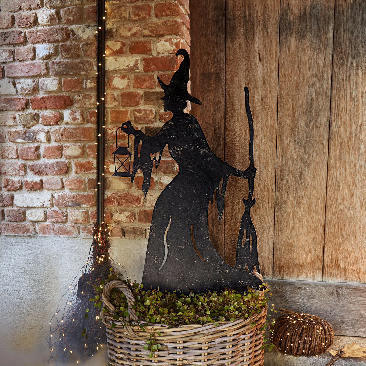 Asta da giardino Talmora Decorazione in metallo nero a forma di strega con lanterna e scopa accanto a un cesto di vimini con piante