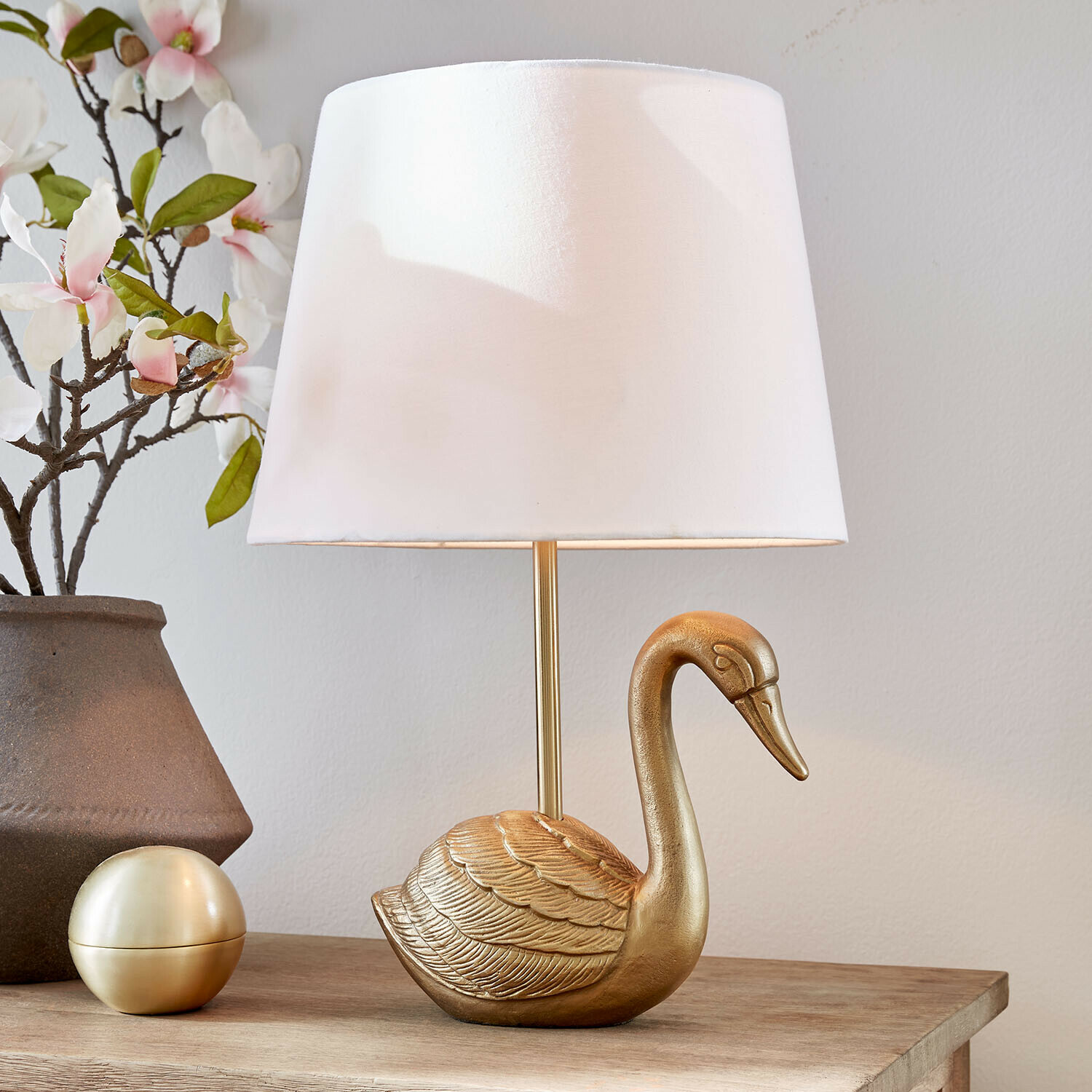 Lampada da tavolo Élégance Dorée Lampada da tavolo dorata a forma di cigno con paralume bianco su tavolo in legno con decorazioni floreali