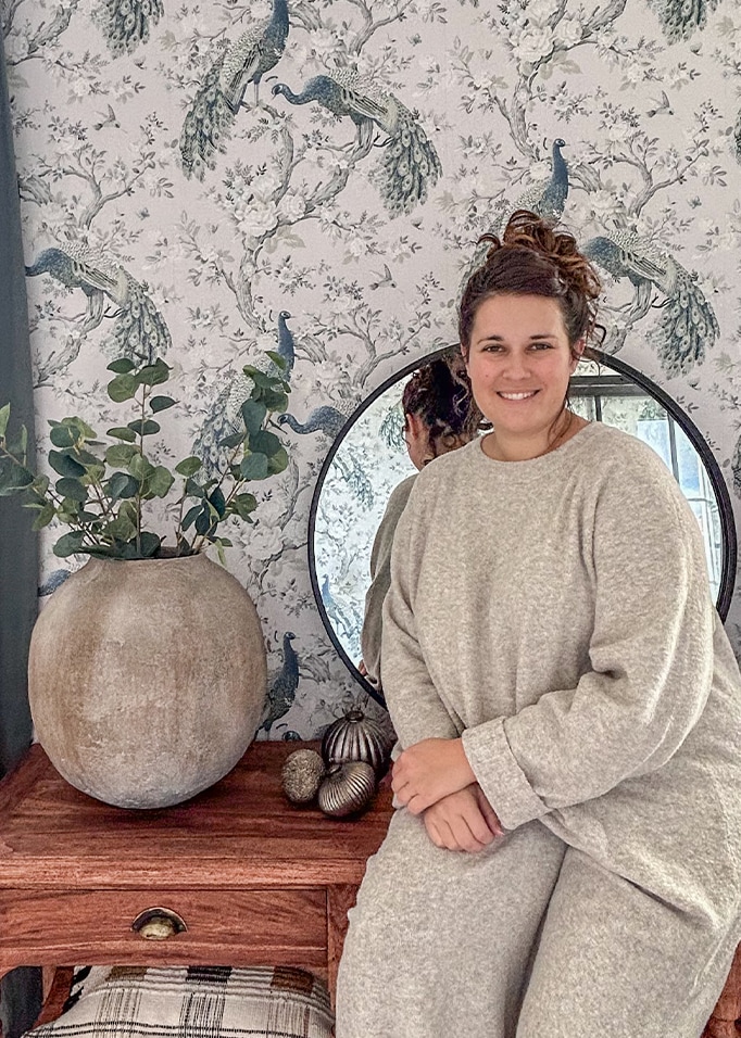 Donna con maglione grigio seduta vicino a tavolo in legno con grande vaso in ceramica e specchio davanti a carta da parati con pavone